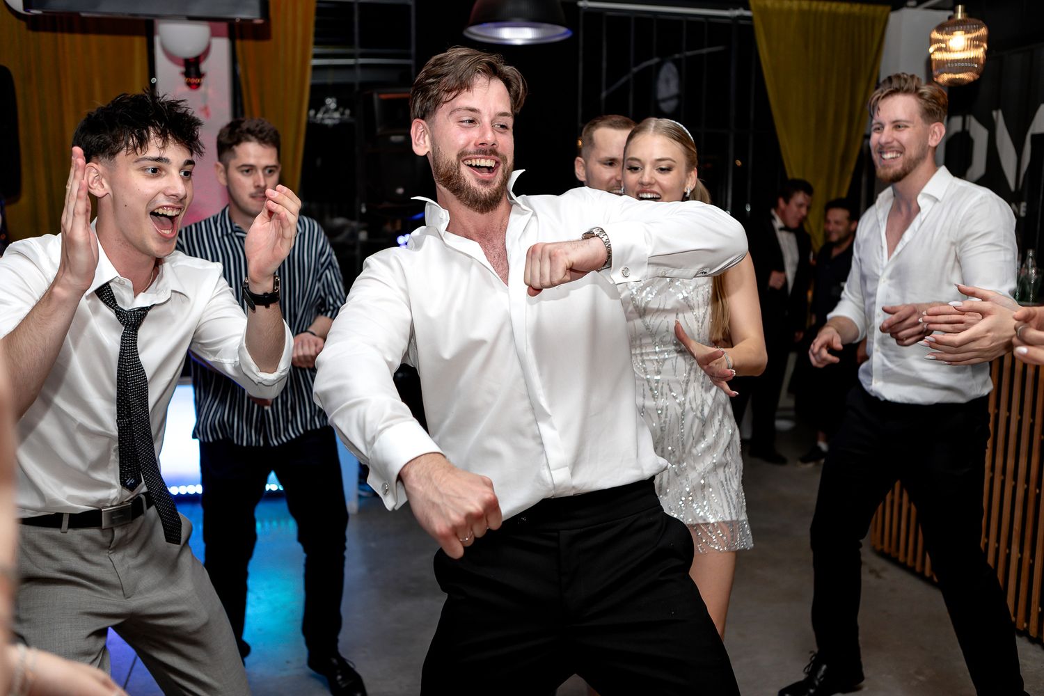 Party guests dancing and celebrating in formal attire at an indoor event.
