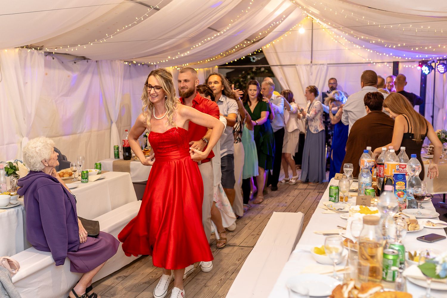 Wedding guests dance and celebrate under a white tent decorated with draped fabric and soft purple lighting.