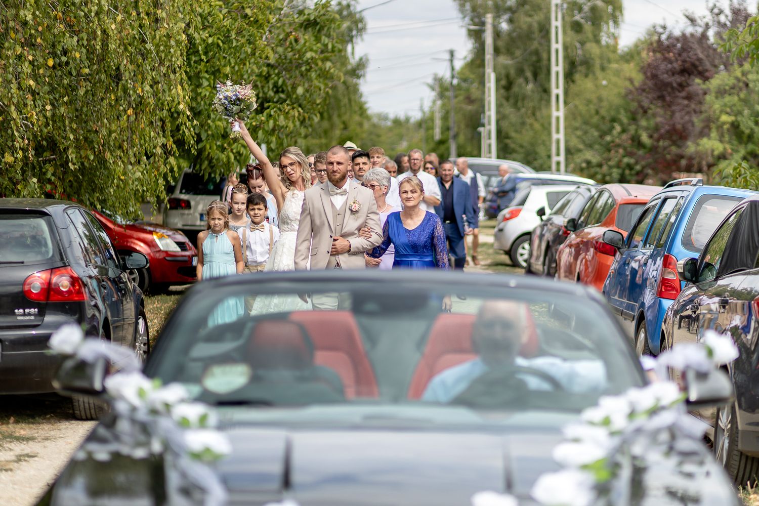 A line of parked cars stretches along a tree-lined path with people walking in the background on a sunny day.