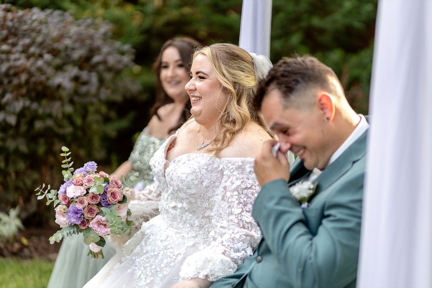 A joyful wedding couple shares an intimate moment together outdoors surrounded by white columns and greenery.