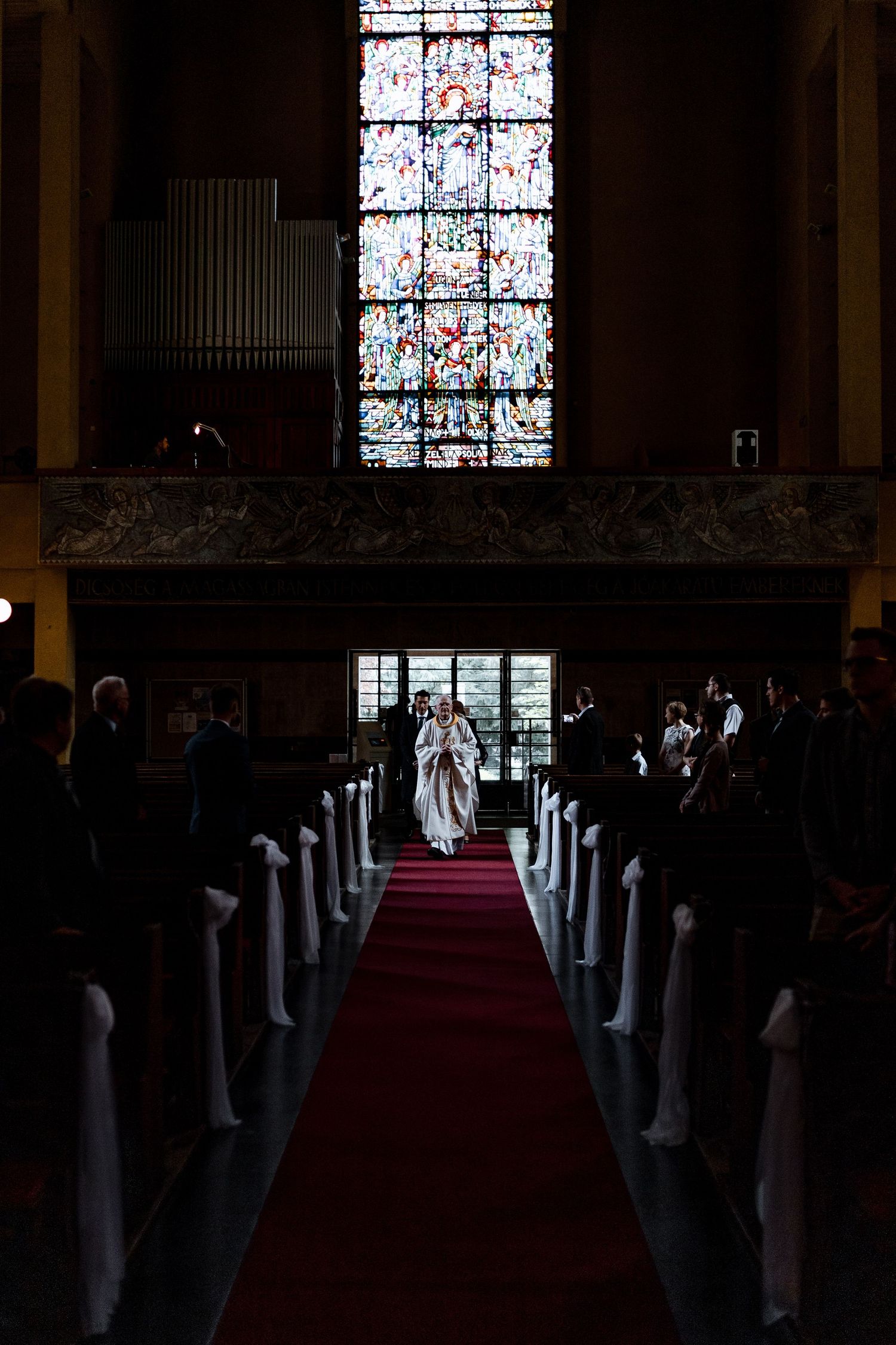 Church aisle with red carpet runner and white decorative elements leading to altar with stained glass windows.