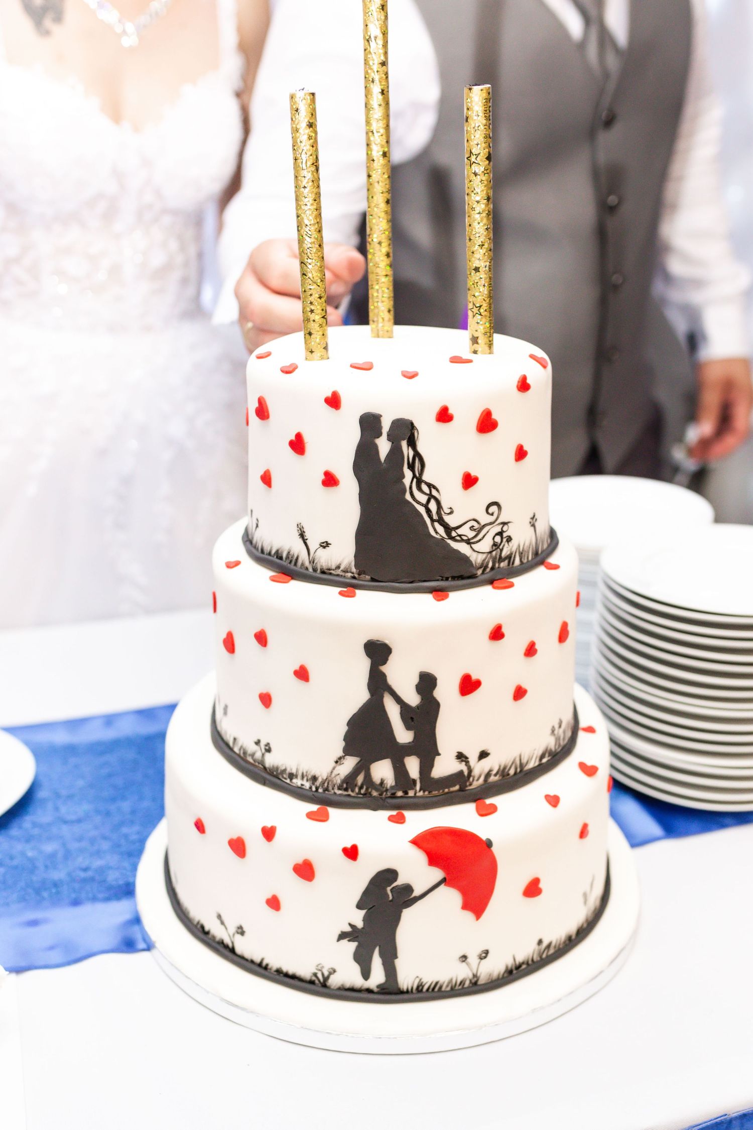 A three-tiered wedding cake decorated with silhouettes of a couple and red hearts against a white background.