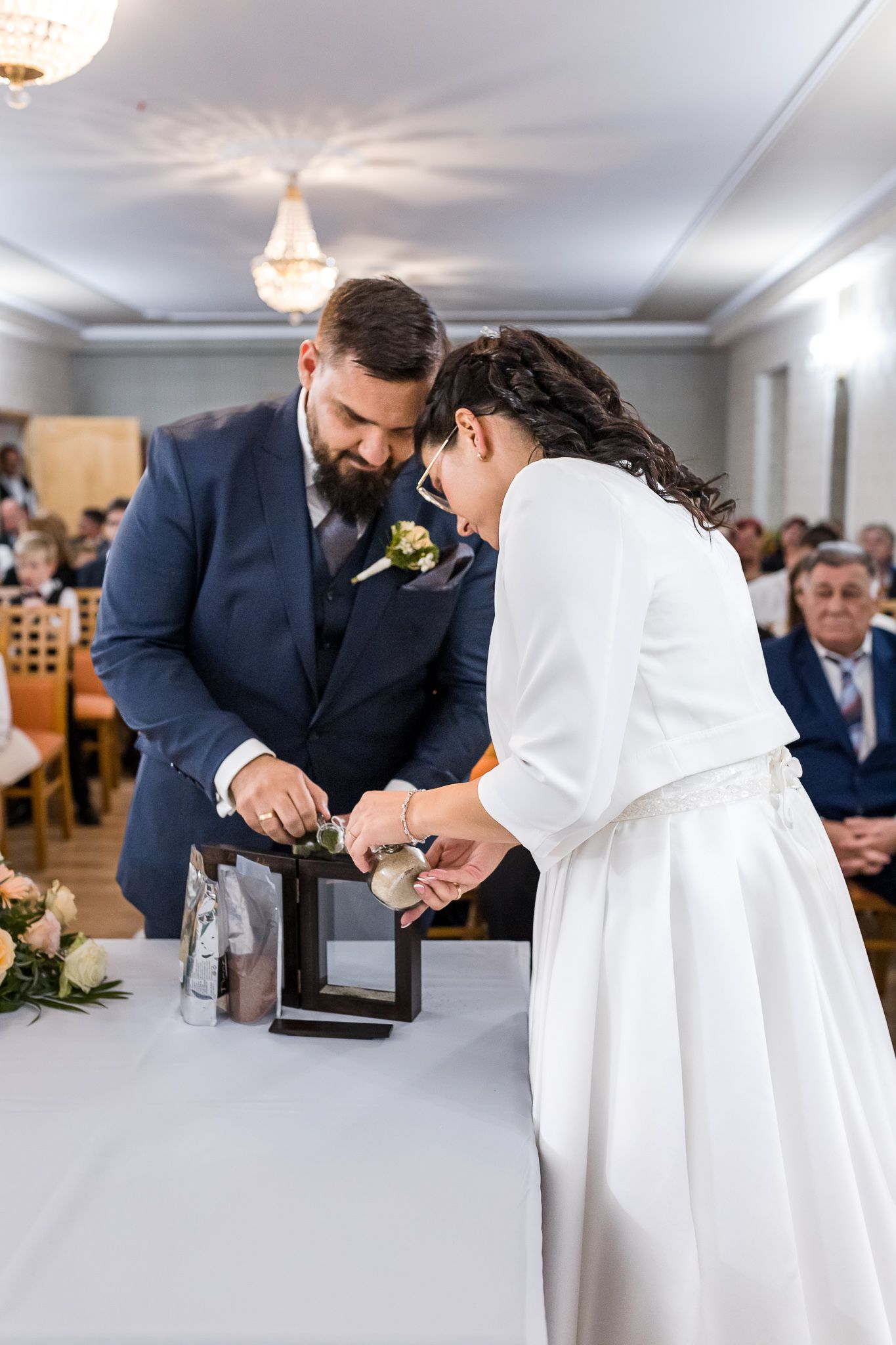 A couple in wedding attire pours sand together during their unity ceremony while guests watch in an elegant venue.