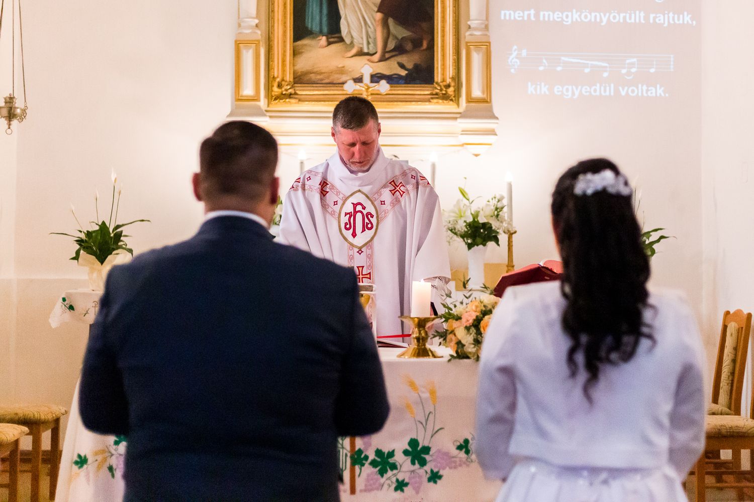 A Catholic priest in white vestments leads a religious ceremony at an ornately decorated altar in a church.
