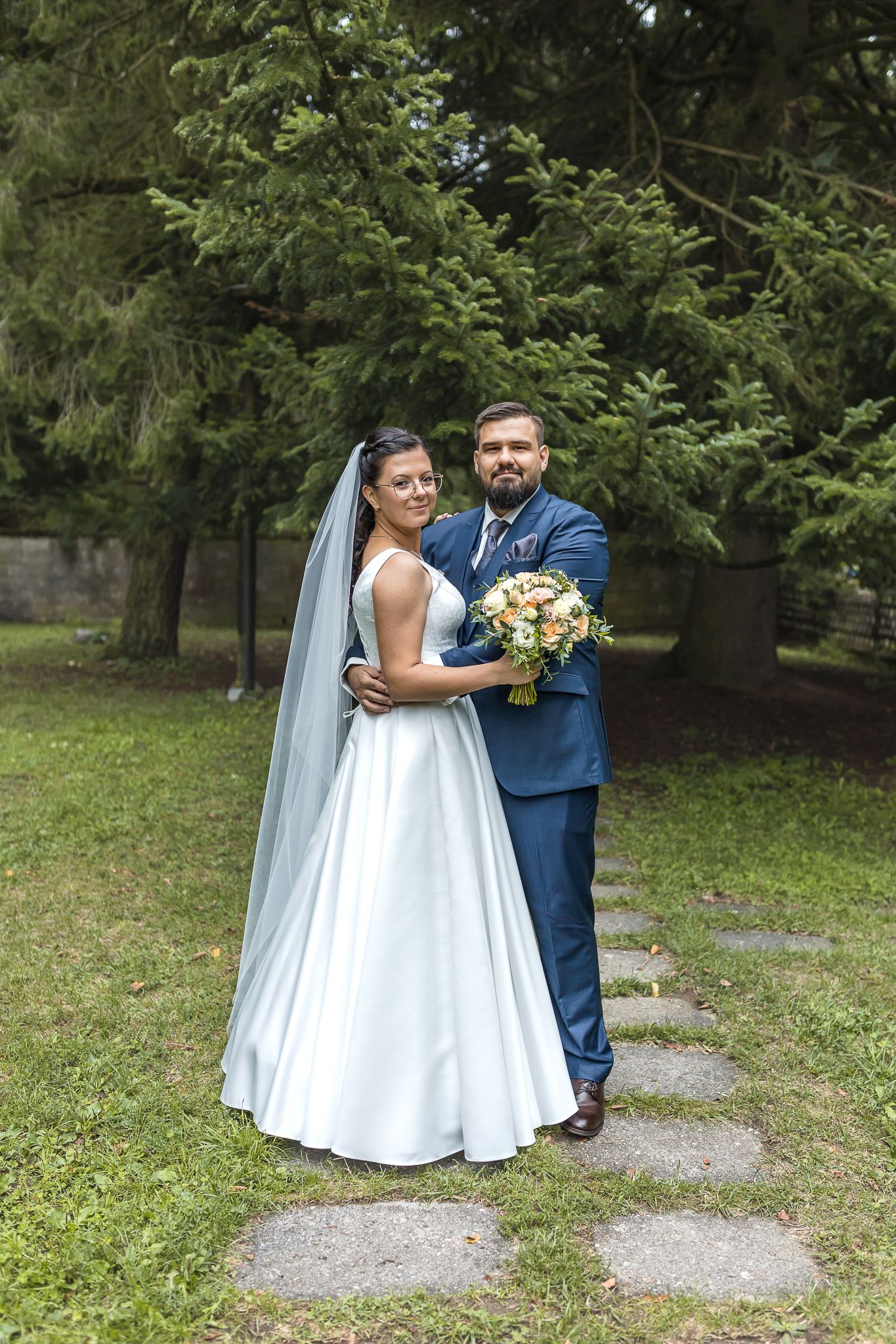 A newlywed couple poses outdoors in a garden setting with the bride in white and groom in blue holding a floral bouquet.