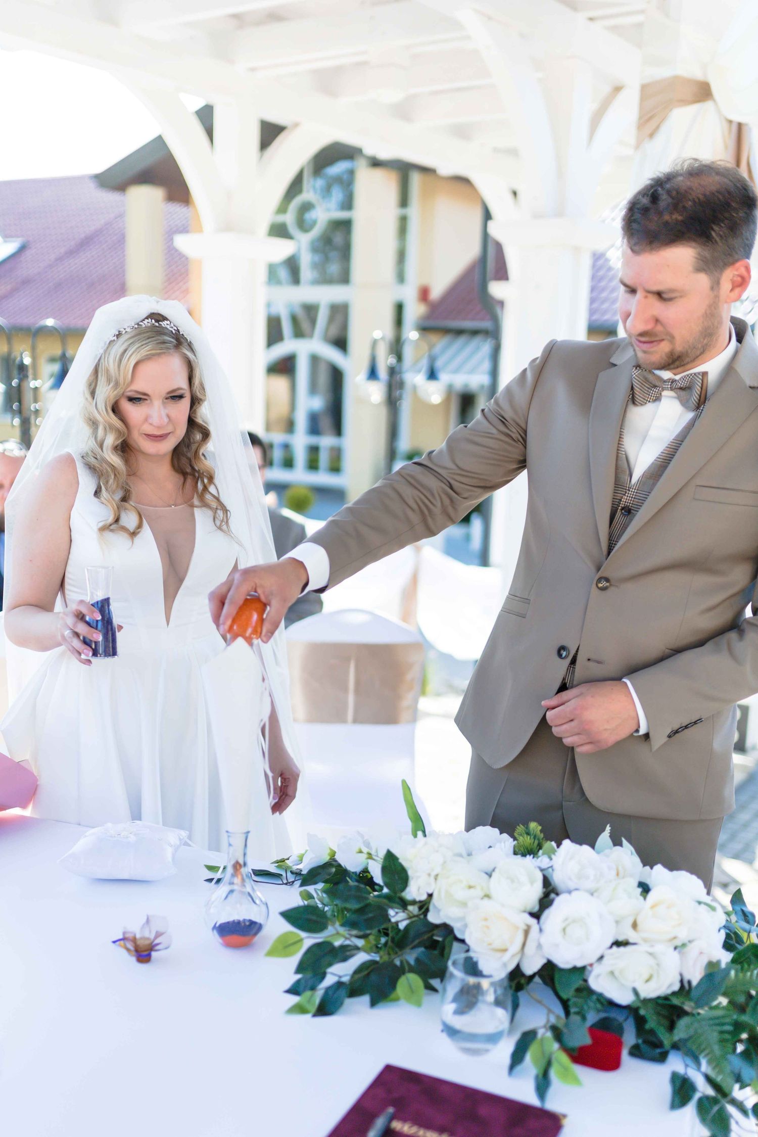 Couple in formal attire sits at an elegant table with white floral arrangements at an outdoor wedding reception.