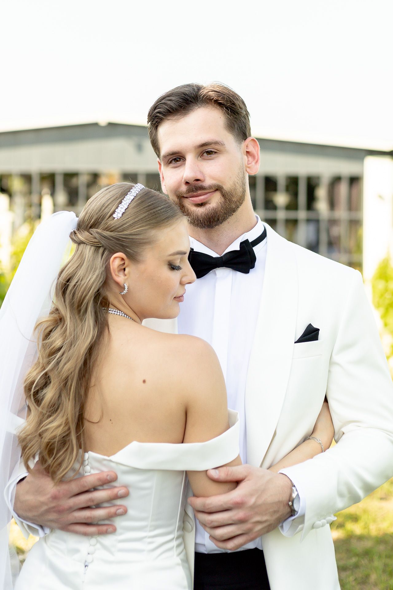 Bride in strapless gown and groom in white tuxedo share tender embrace during outdoor wedding.