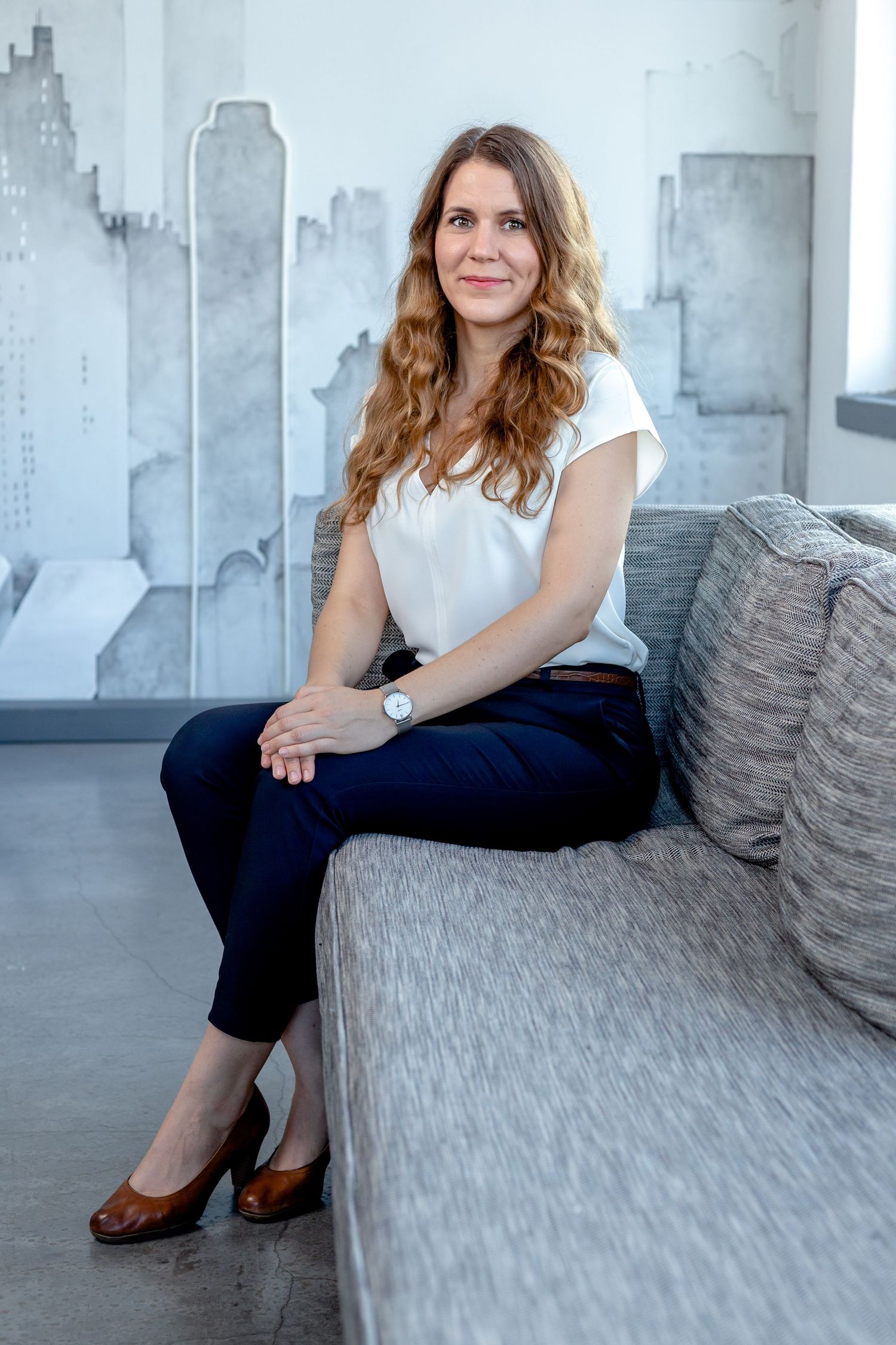 Professional seated on grey textured couch wearing white top and navy pants in industrial space.