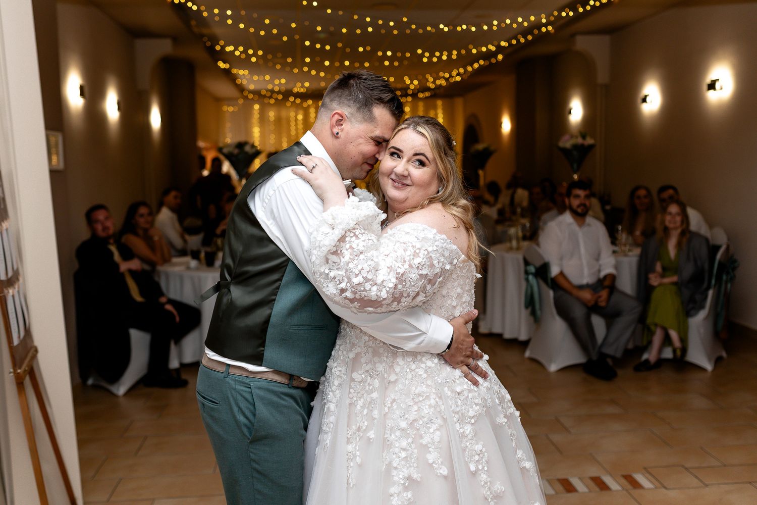 A couple shares a romantic dance moment under twinkling string lights at an elegant indoor wedding reception.