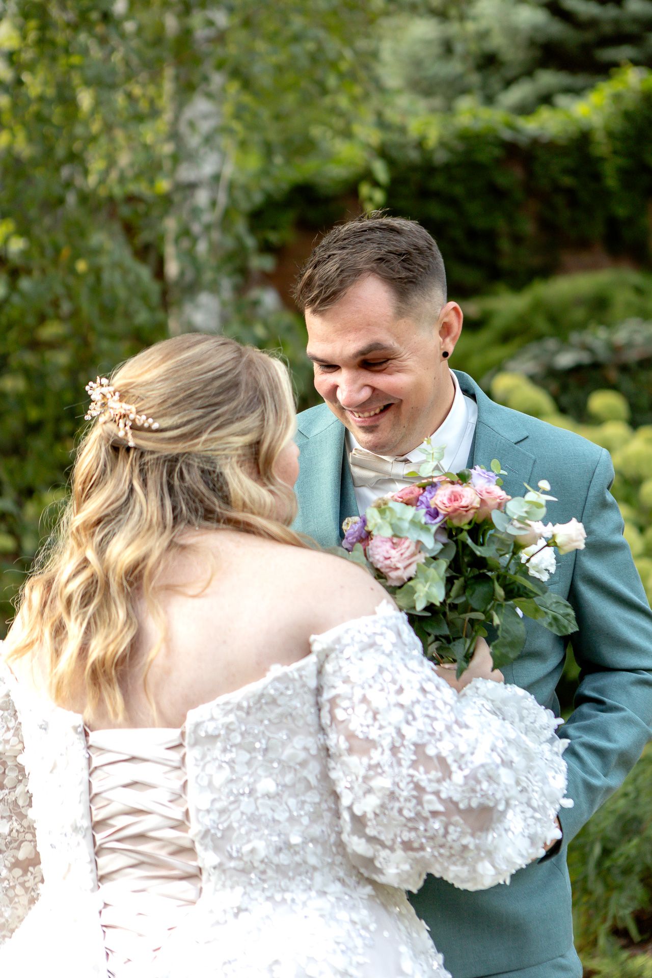 Couple in teal suit and lace wedding dress embrace during outdoor garden photoshoot.
