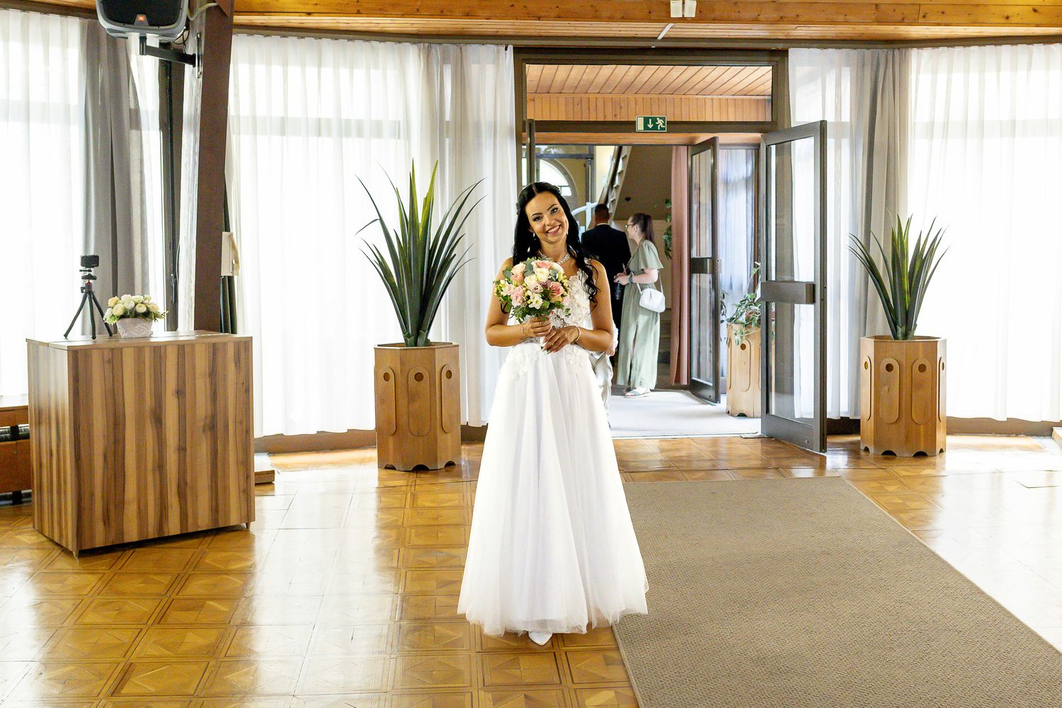 Modern event space with wooden floors and white cocktail tables arranged along a bright windowed wall.
