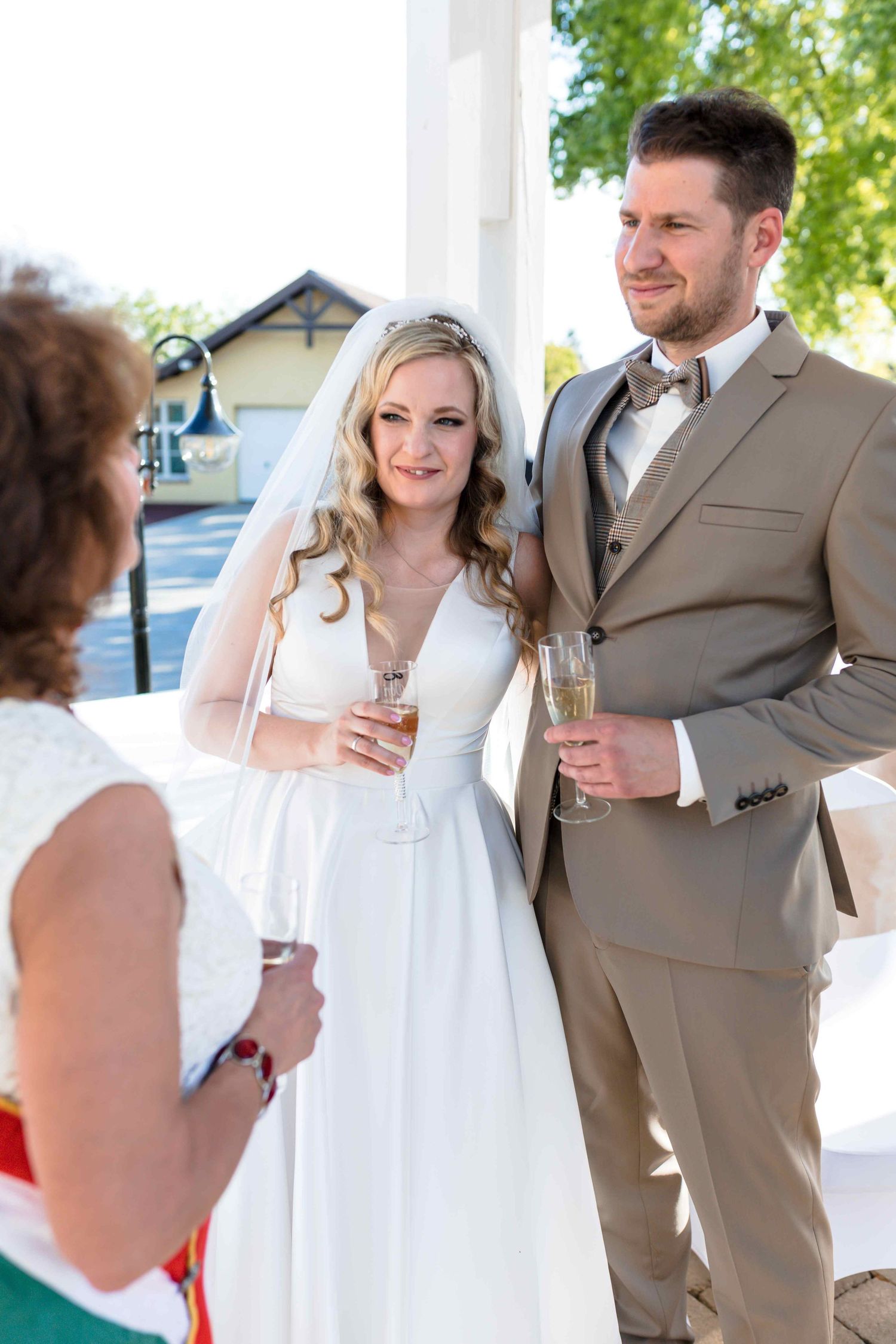 Wedding couple in tan suit and white dress share champagne toasts during outdoor celebration.