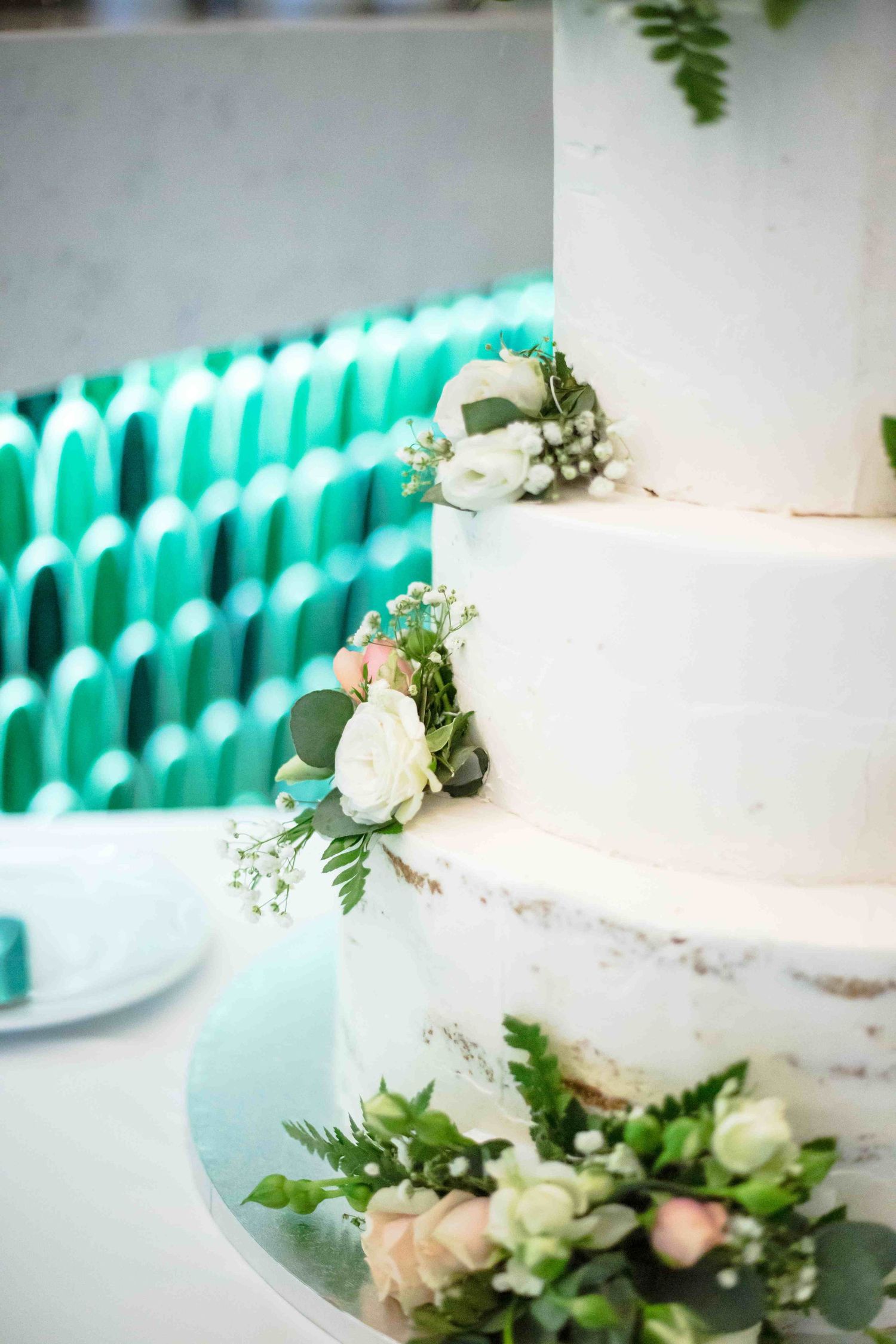 White wedding cake decorated with fresh roses and greenery against a turquoise backdrop creates an elegant display.