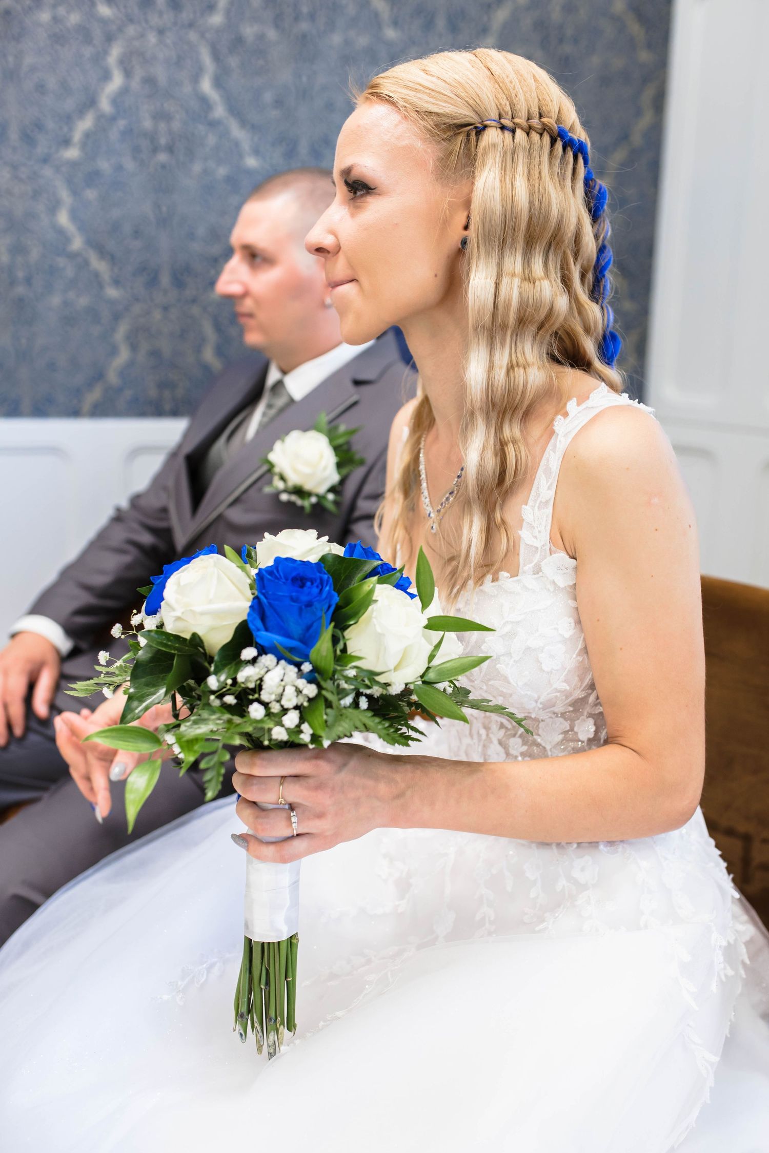 Wedding couple in formal attire with a bouquet featuring blue and white roses against a textured wall.