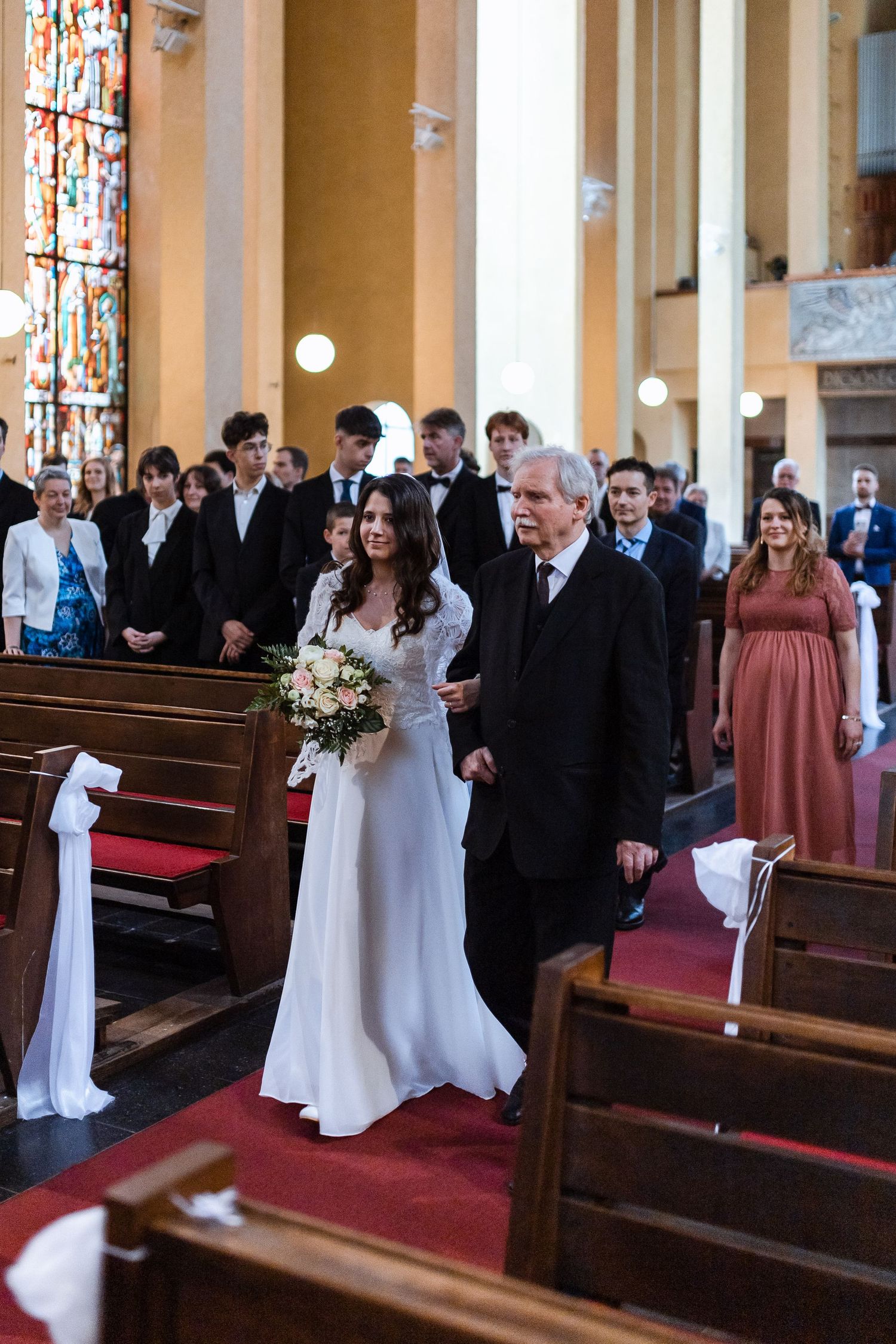 A wedding ceremony inside a church with stained glass windows, where a couple walks down the red-carpeted aisle.