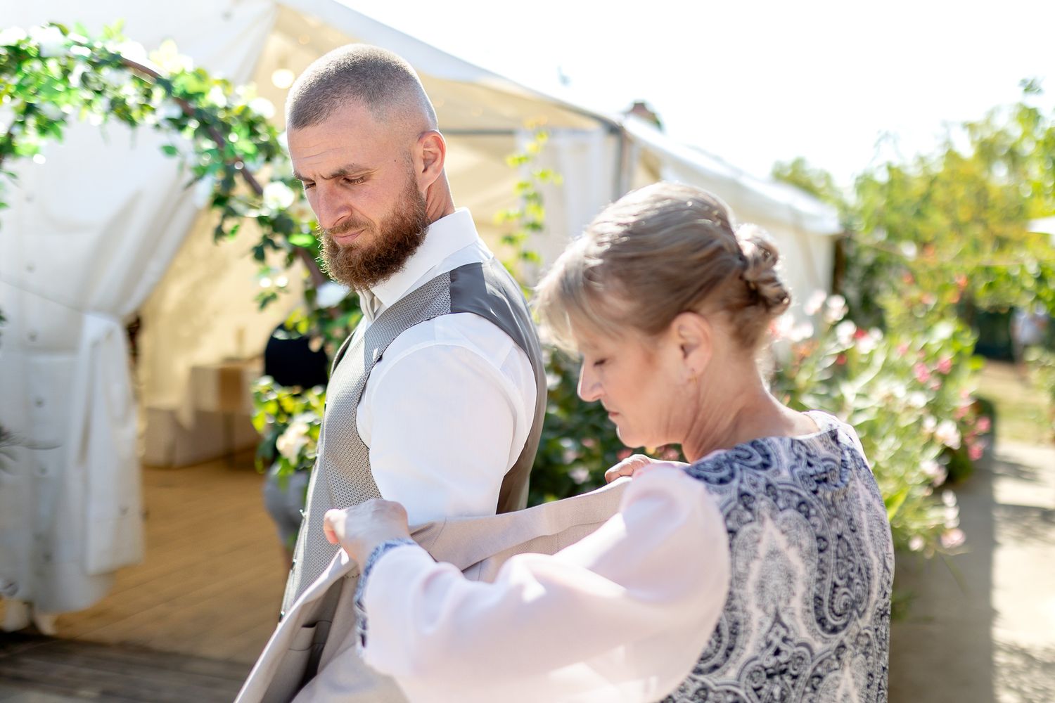 Two people stand together outdoors, one wearing a formal vest while the other adjusts their attire in bright sunlight.
