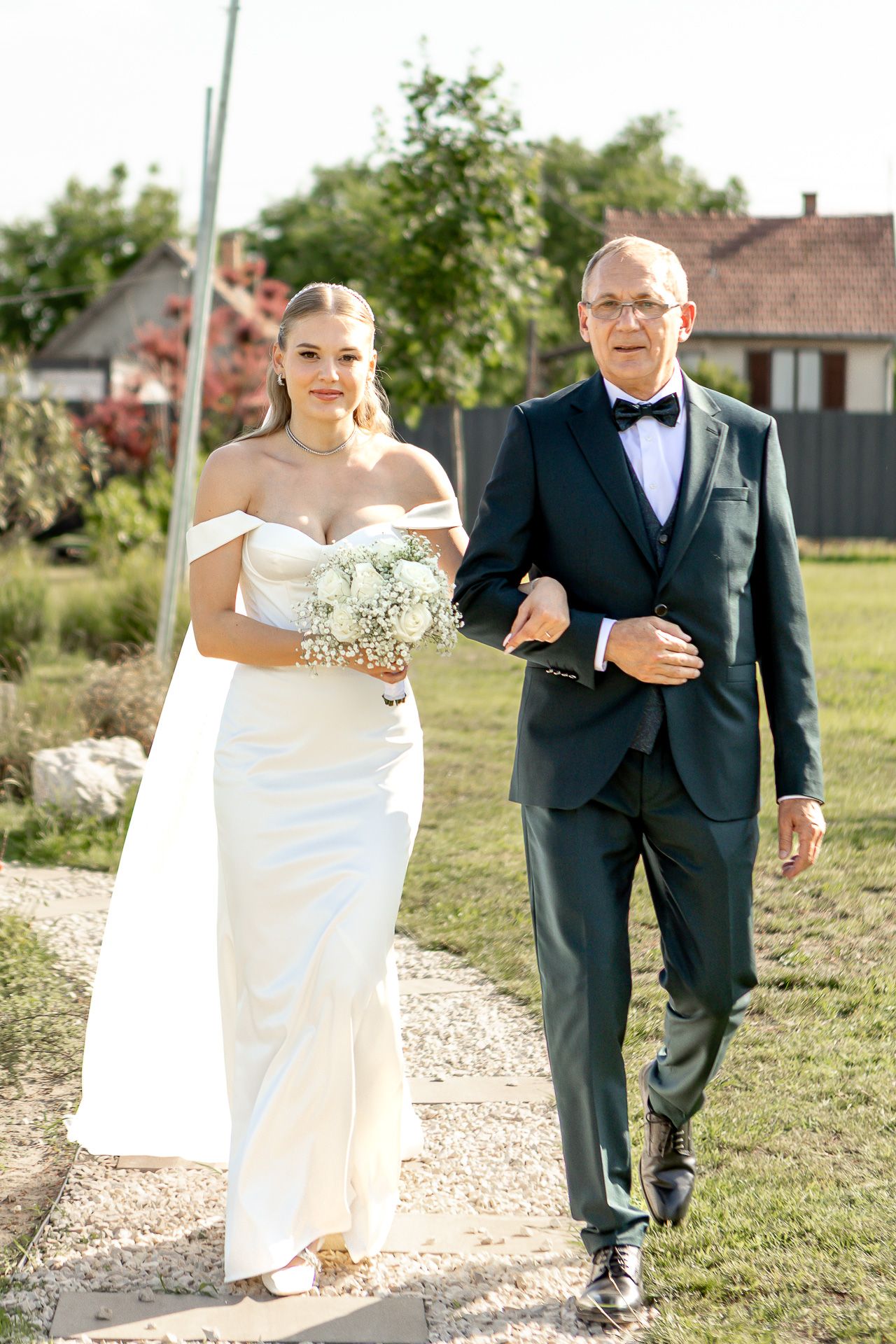 Bride in white gown walks down gravel path escorted during outdoor ceremony.