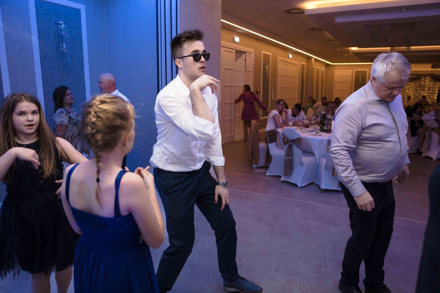 People dancing and socializing at an indoor event with ambient lighting and white chairs in the background.