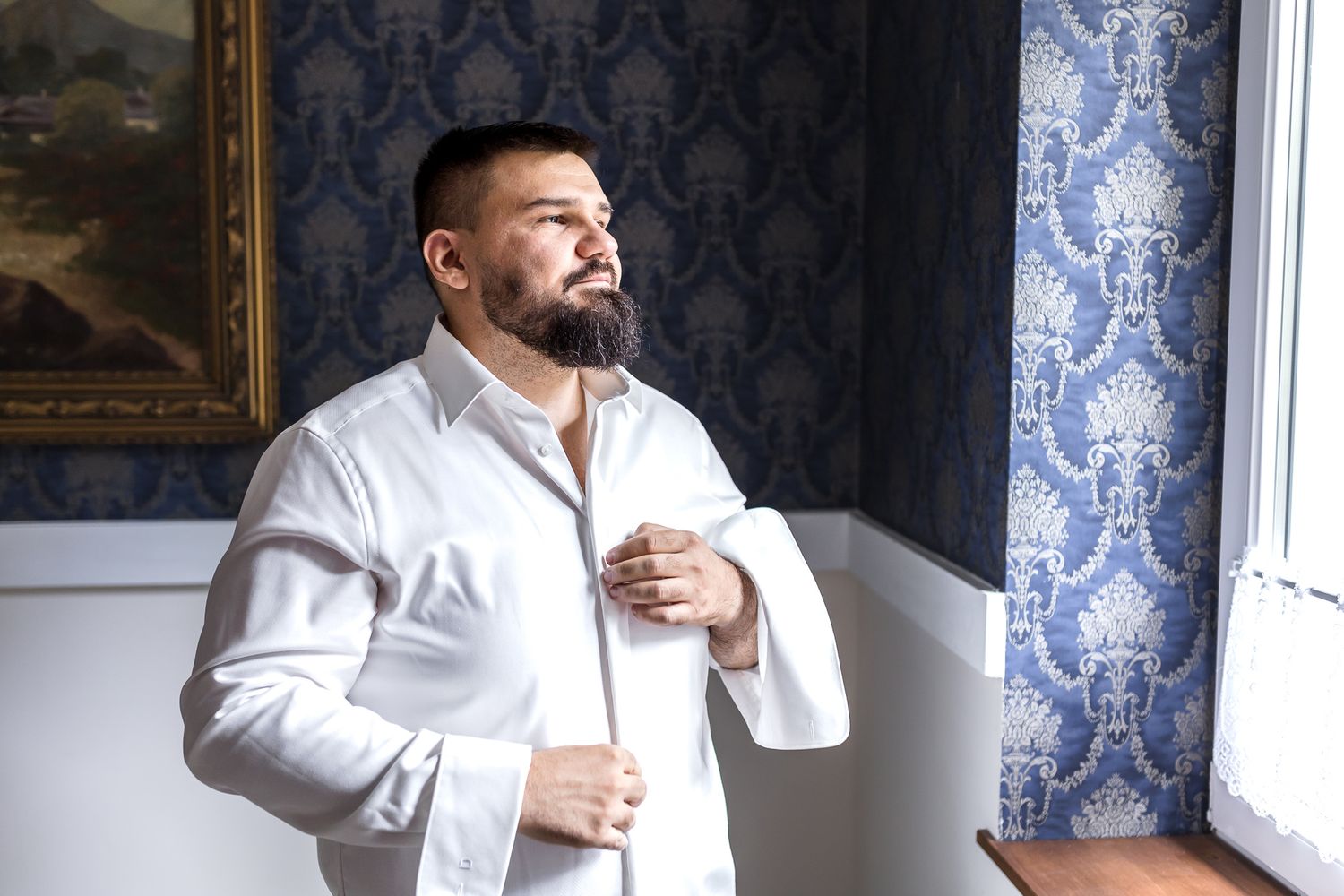A person in a white shirt stands near a window with blue patterned wallpaper in a contemplative pose.