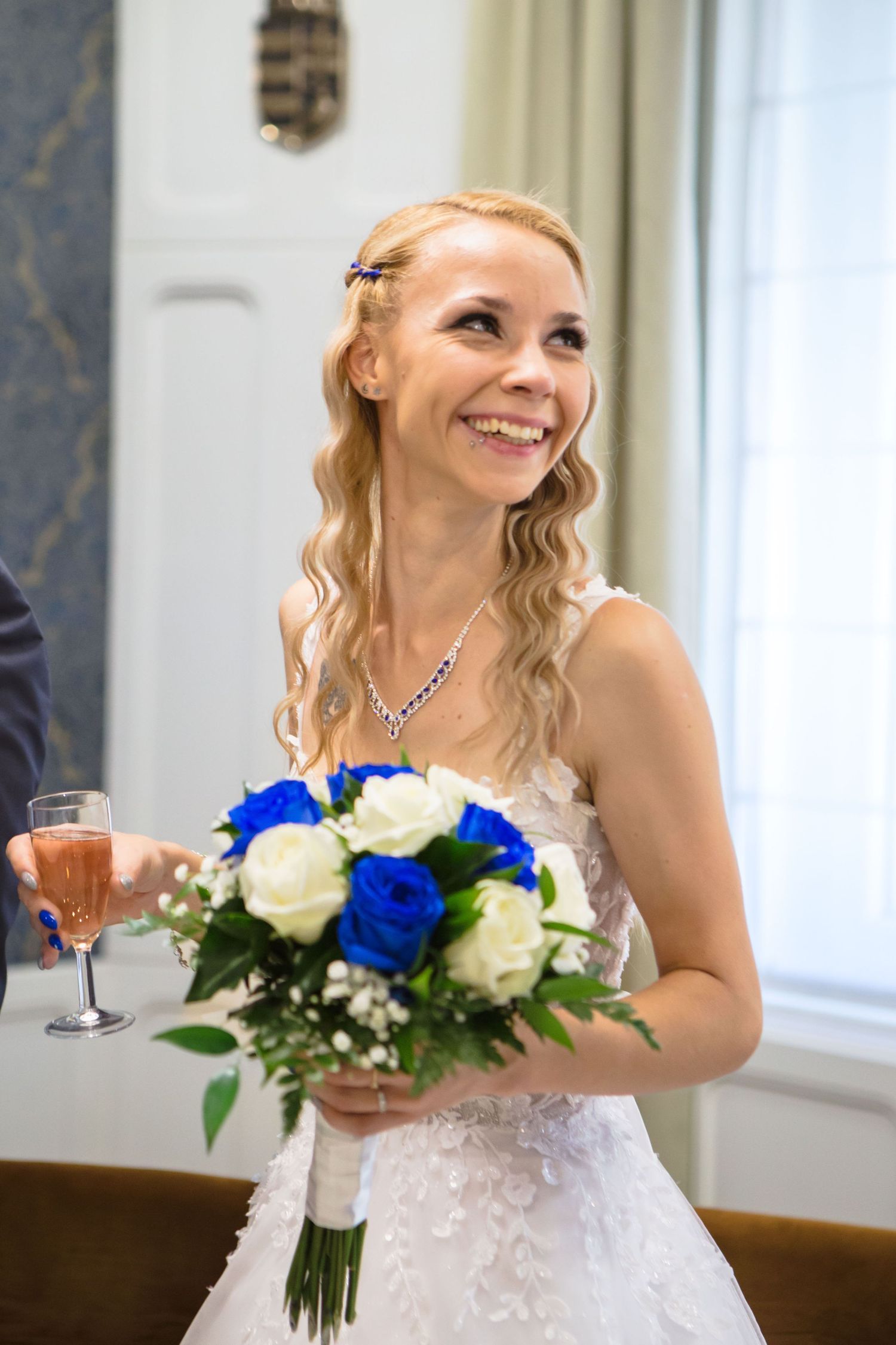 A bride in a white lace dress smiles while holding a blue and white rose bouquet at her wedding celebration.