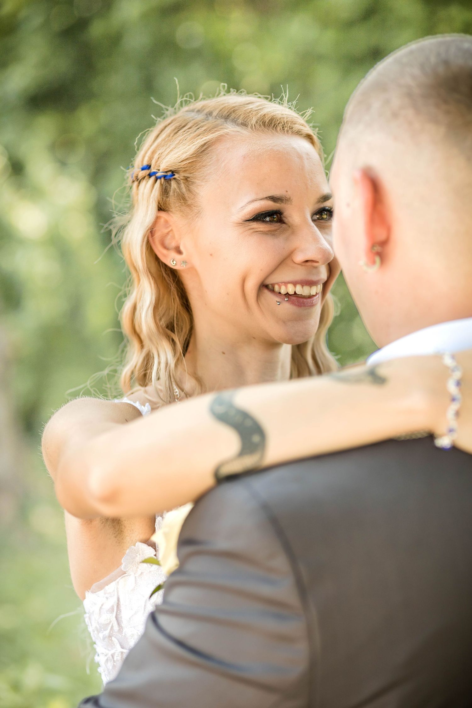 A romantic wedding couple embraces outdoors in a natural setting with soft sunlit bokeh in the background.