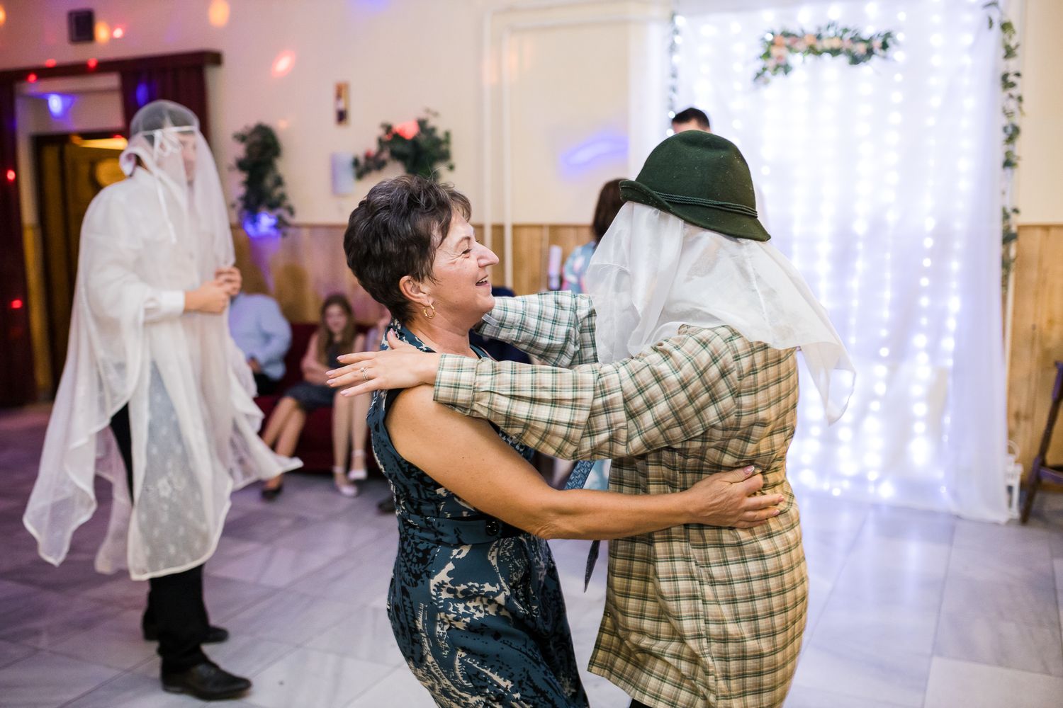 Couples dance together during a wedding reception in a brightly lit ballroom with white curtains and decorative lighting.