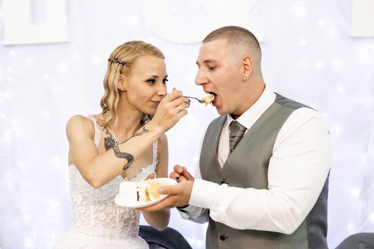 Couple in wedding attire share a piece of cake during a reception celebration with white decor in the background.