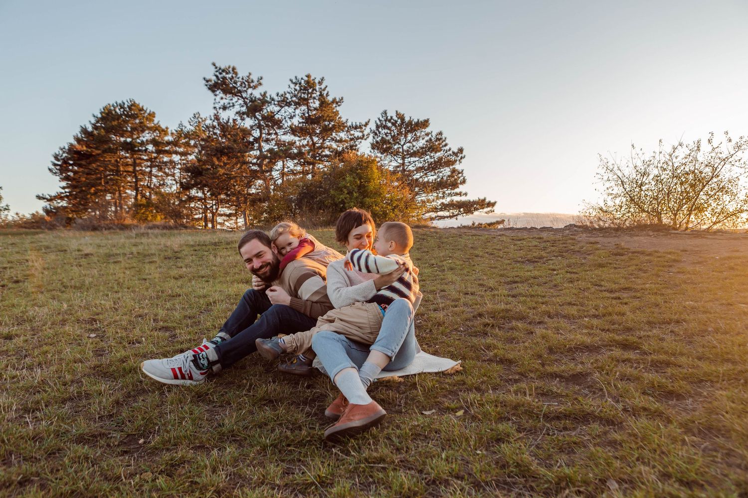Family relaxing together on a grassy slope during sunset with pine trees in background.