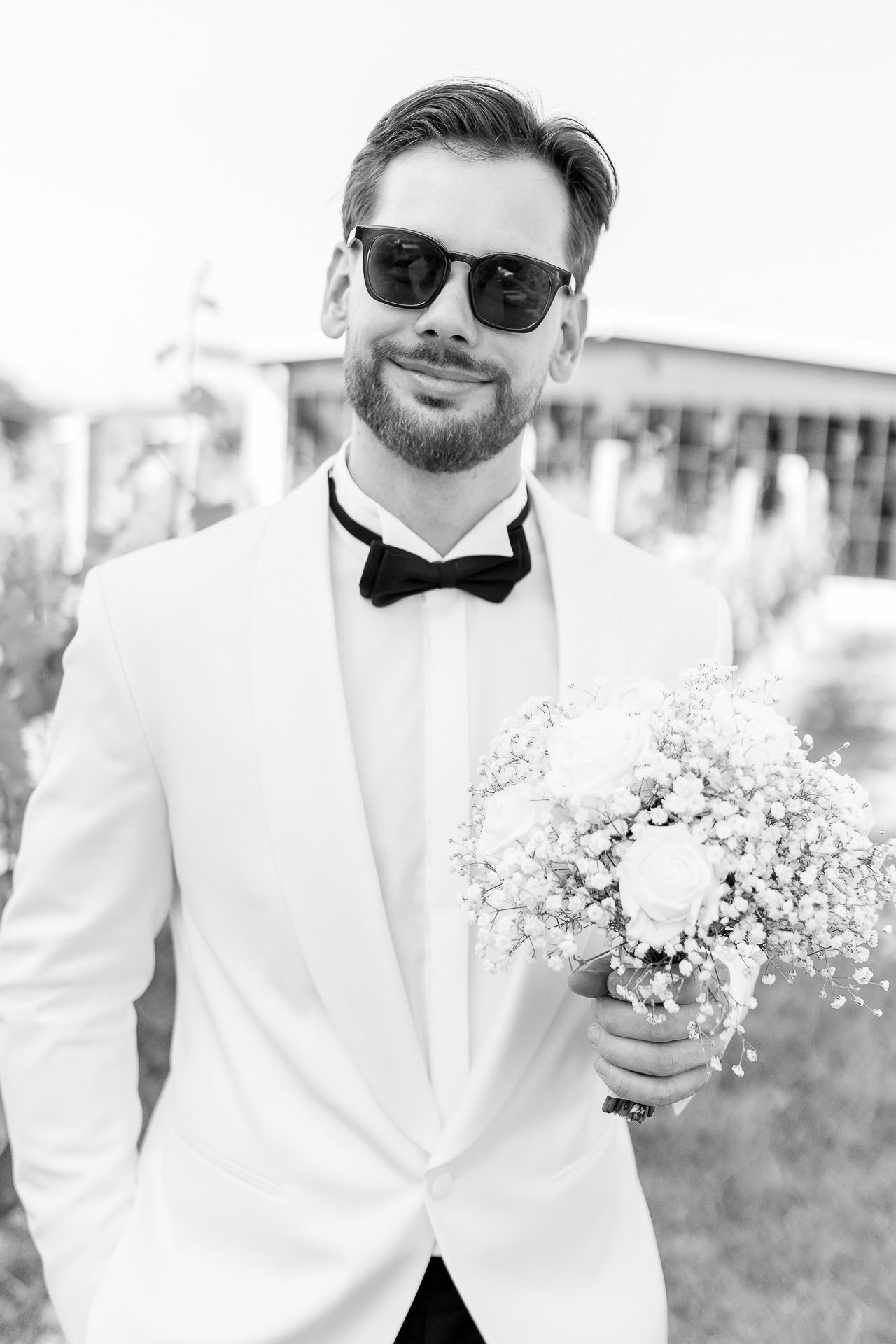 Stylish groom in white tuxedo and sunglasses holds delicate baby's breath bouquet in black and white photo series.