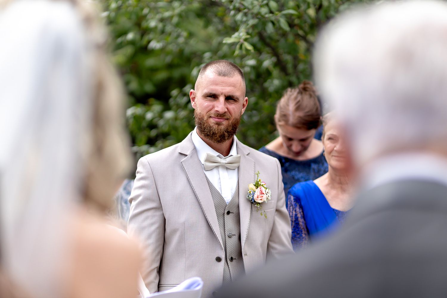 A groom in a light gray suit with bow tie and boutonniere stands at attention during an outdoor wedding ceremony.