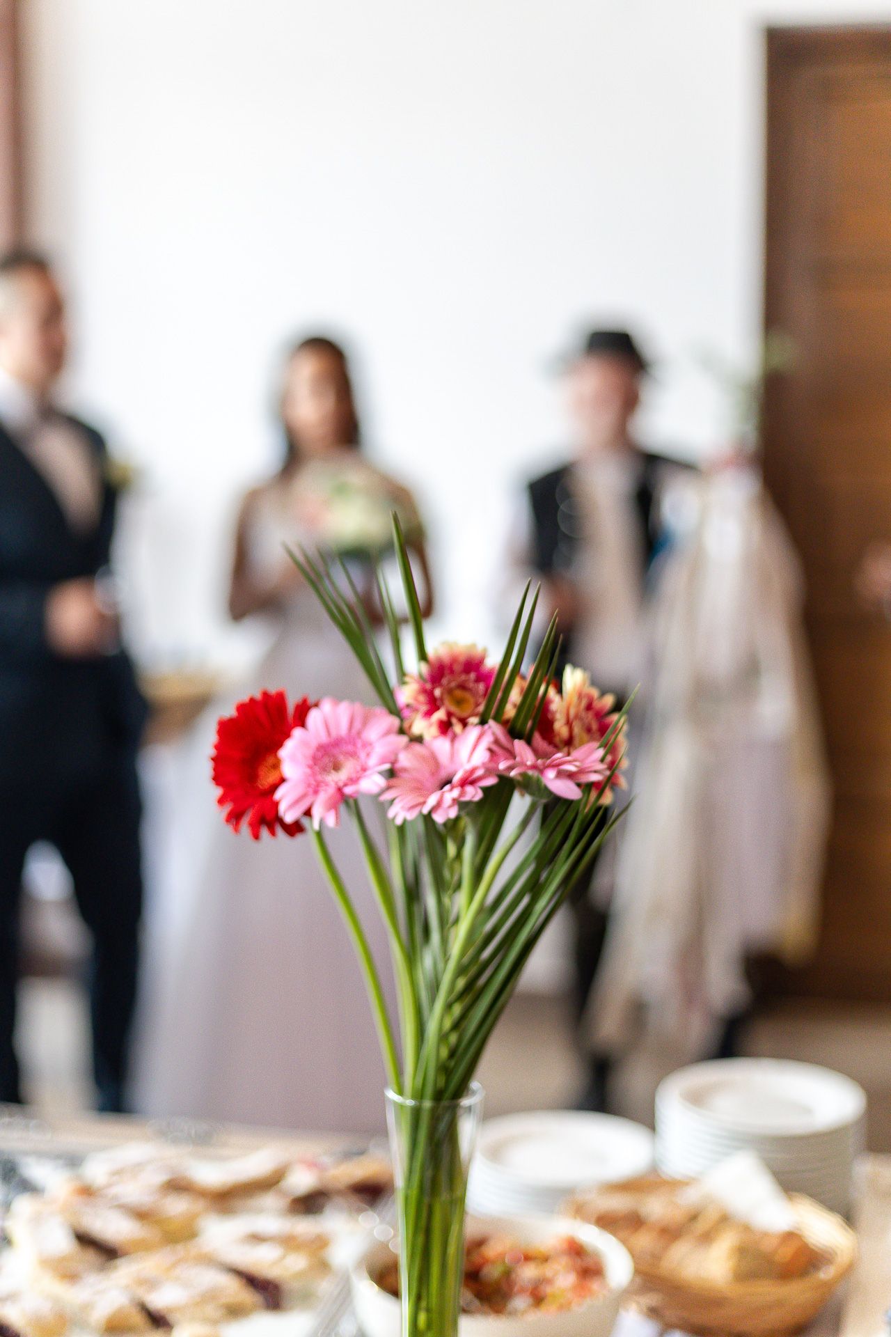 Pink and red gerbera daisies in a vase decorate a buffet table at an indoor event with guests mingling in background.