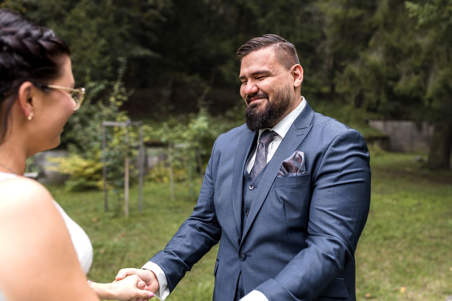 A couple in formal wedding attire shares an intimate moment while exchanging vows in a sunny outdoor garden setting.