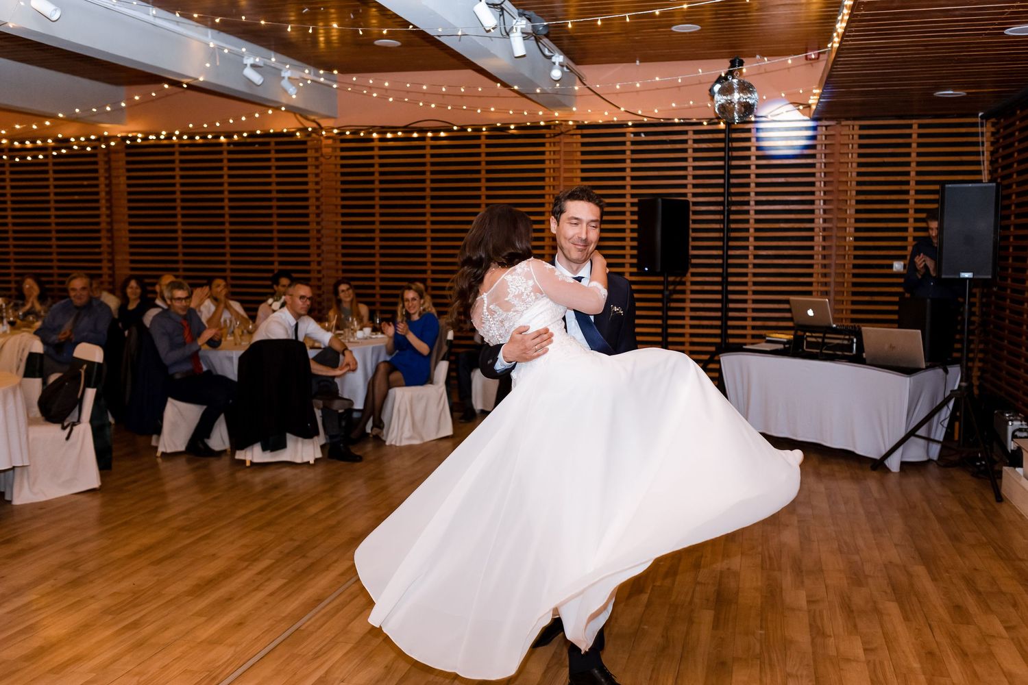 A couple shares their first dance at a wedding reception in a rustic venue decorated with string lights and white linens.