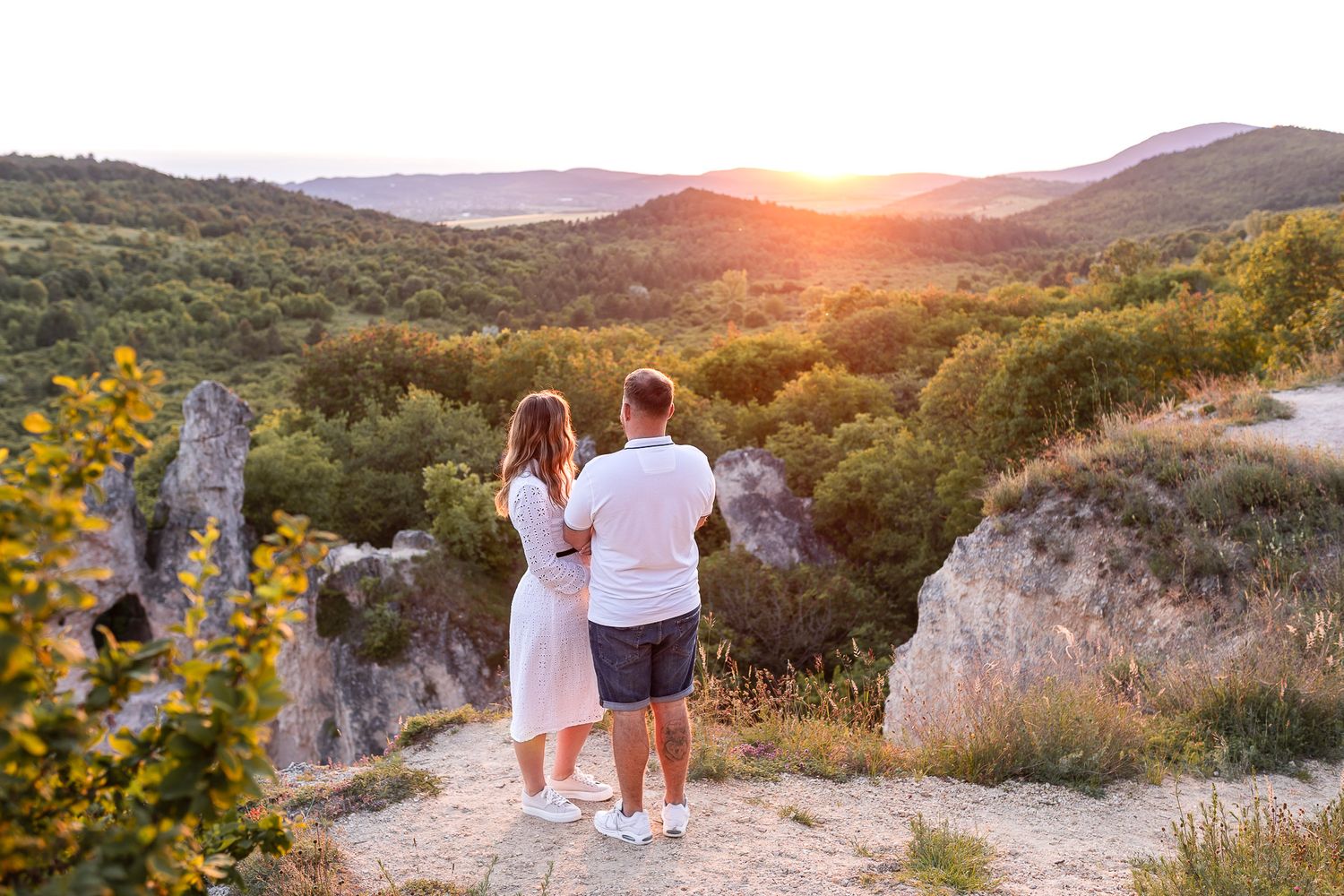A couple stands on a cliff edge admiring a golden sunset over rolling hills and valleys.