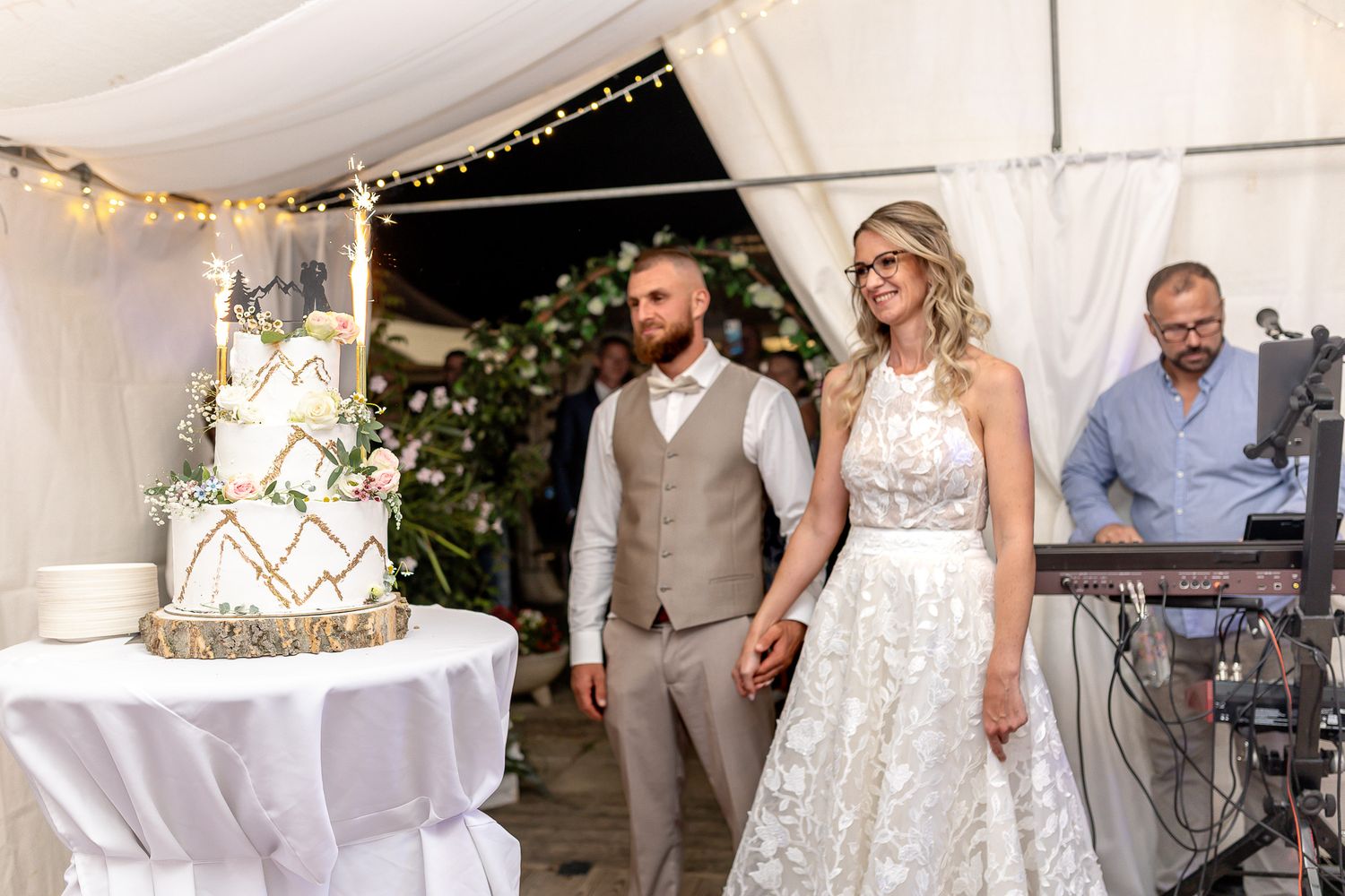 Wedding reception guests dance and celebrate under a white tent decorated with string lights and draping fabric.