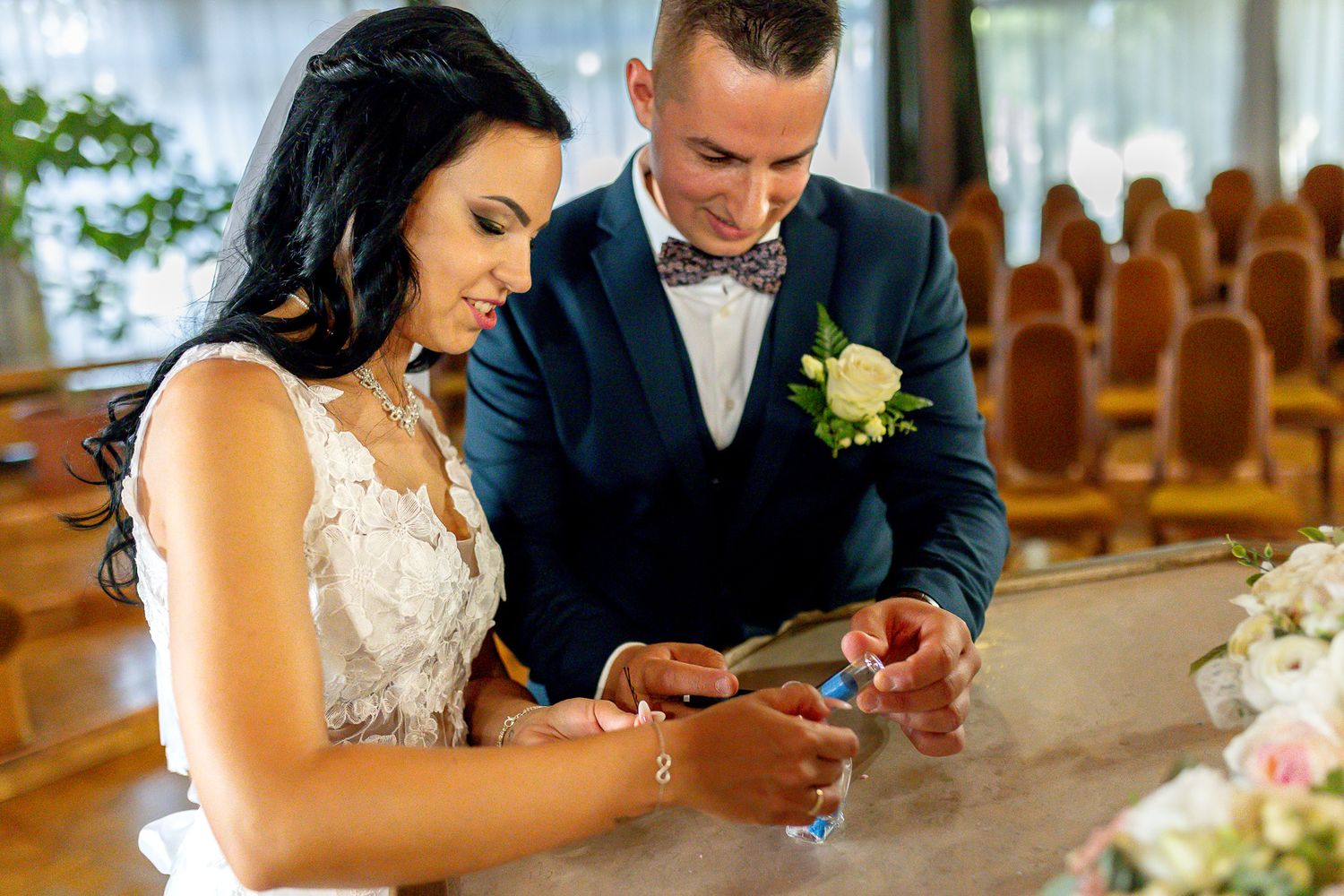 A couple sits together at a wedding reception venue looking at their phones in formal attire.