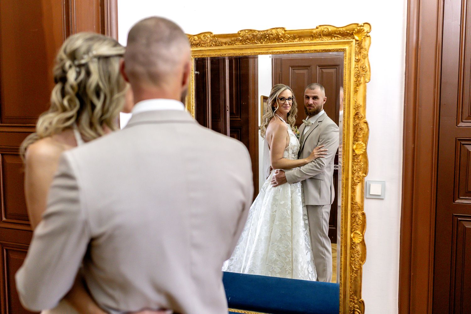 A couple shares an intimate first look moment in an elegant hallway with ornate gold-framed mirrors and wood paneling.