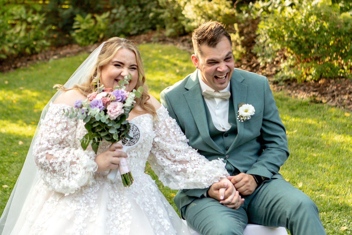 Newlywed couple shares intimate moments while sitting on grass during outdoor wedding photoshoot in garden setting.