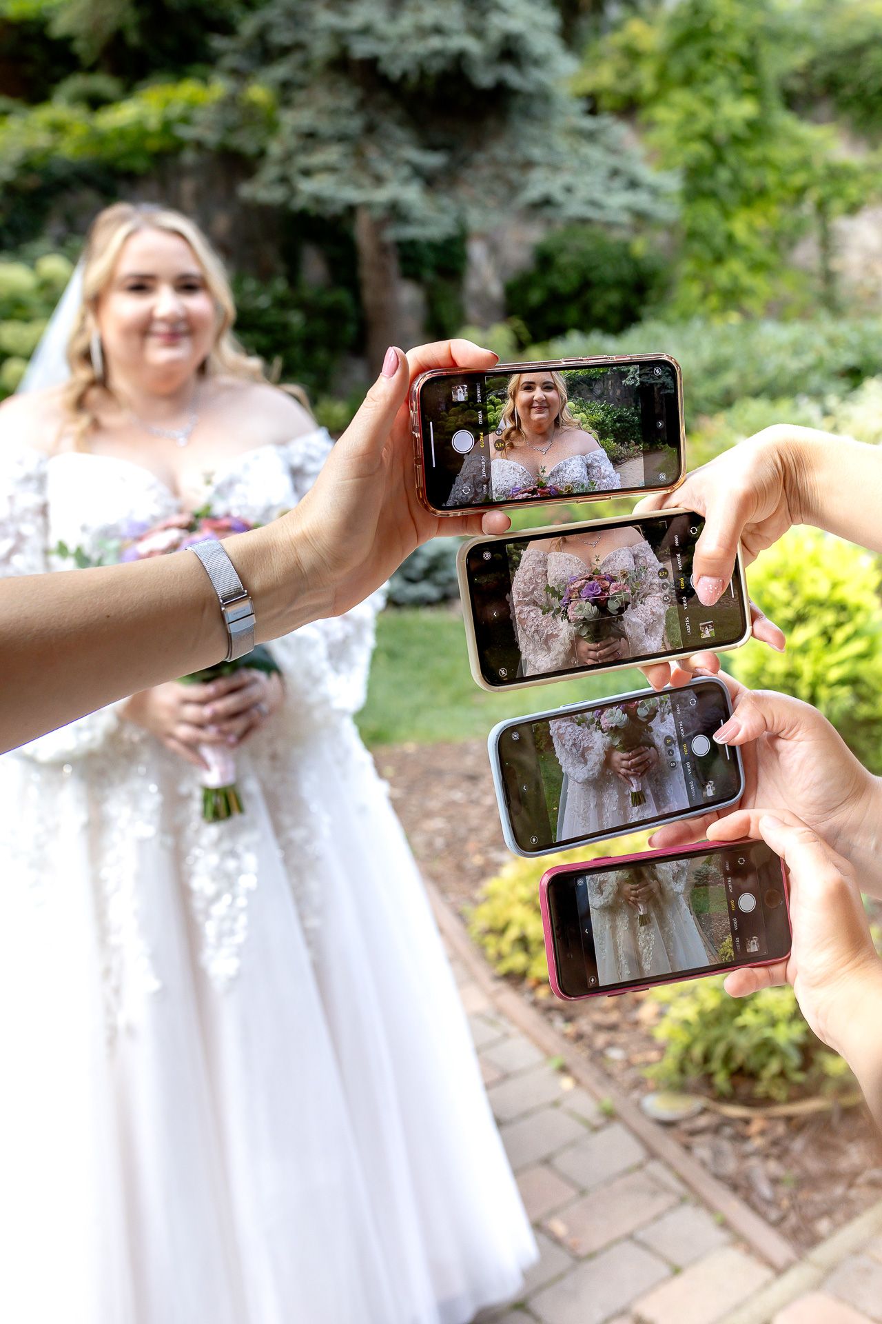 Multiple hands hold phones capturing photos of a person in a white dress on a garden path.
