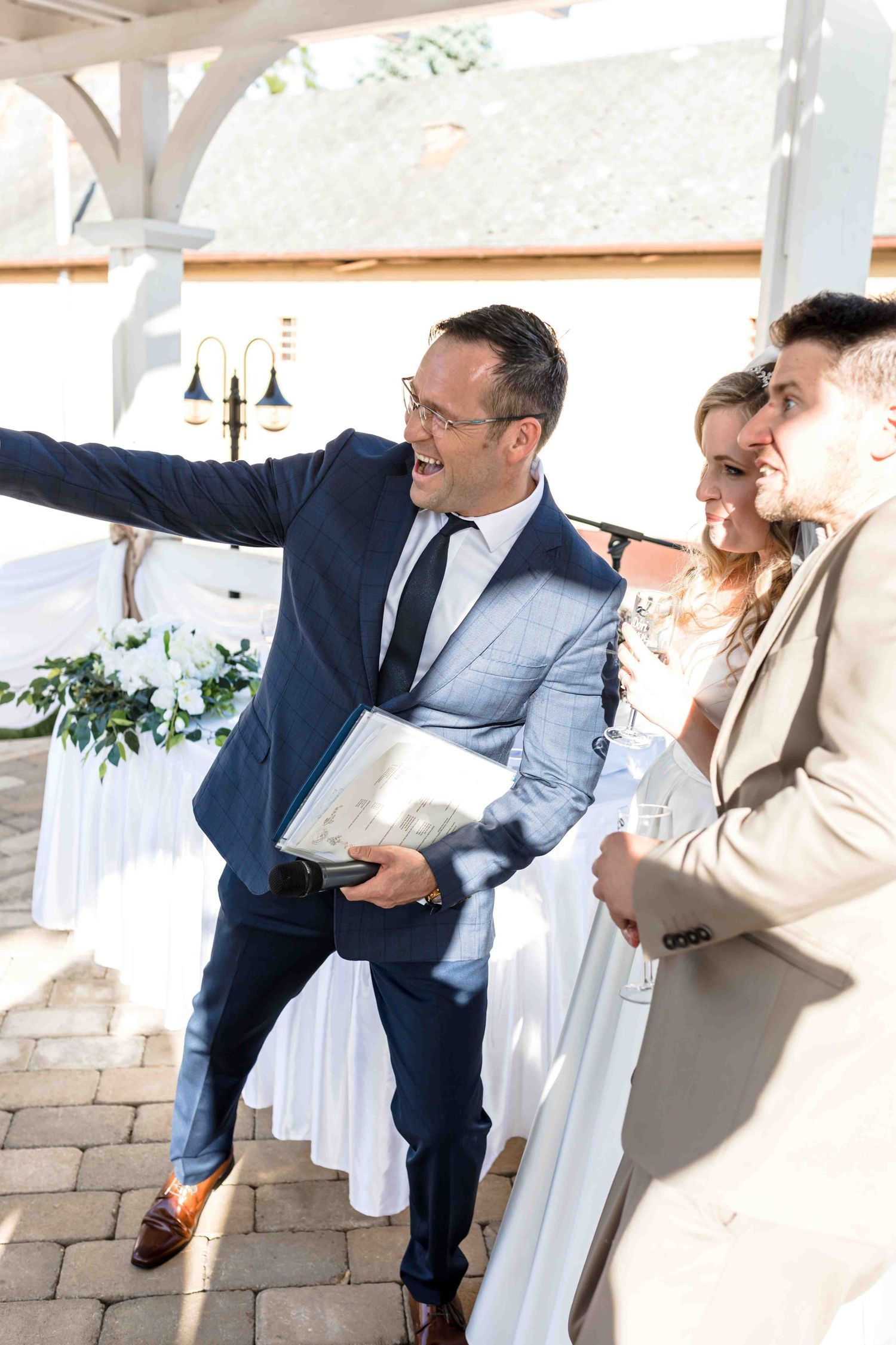 Wedding party laughs together during candid photo session at outdoor venue with stone wall and white decor.