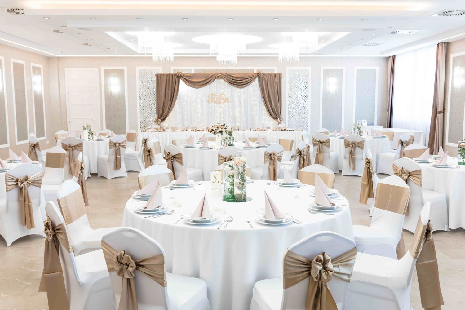 Elegant white wedding reception hall with round tables, cream chair covers, and beige satin bows for a formal event.
