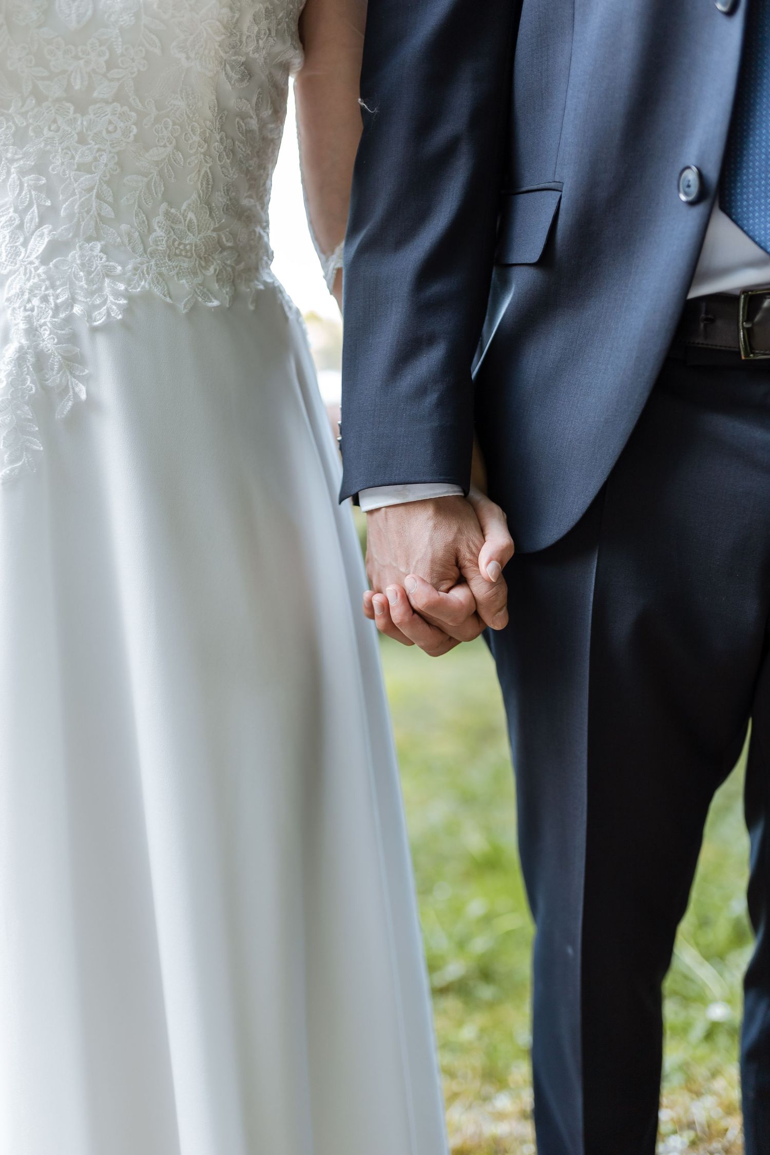 Close-up view of a bride and groom holding hands, showing white wedding dress and navy blue suit details.
