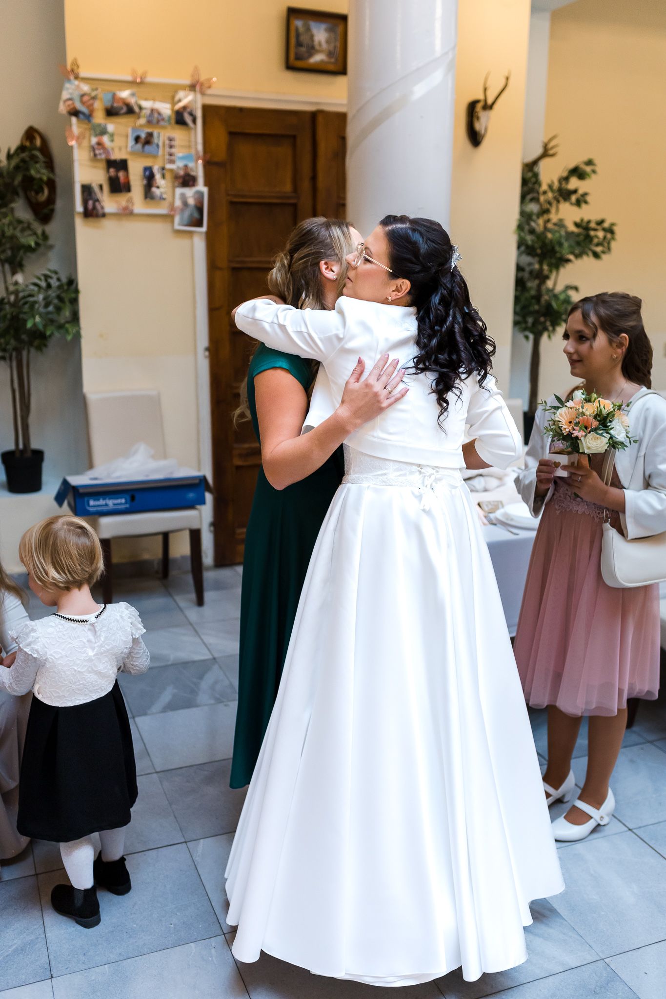 Two people embrace at a wedding celebration while others look on in an indoor venue with tiled floors and decorative walls.