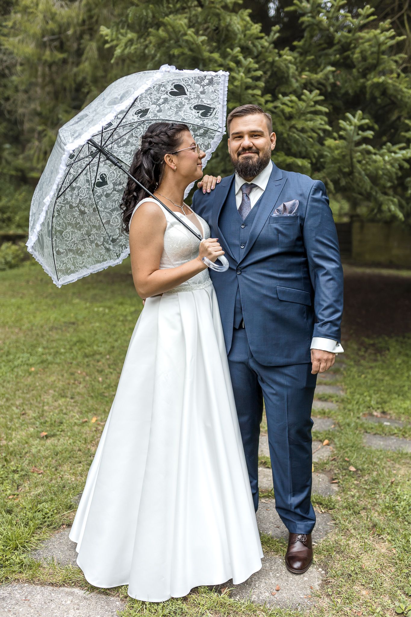 A romantic wedding photo outdoors featuring a white lace parasol, elegant white gown and navy blue suit in a garden setting.