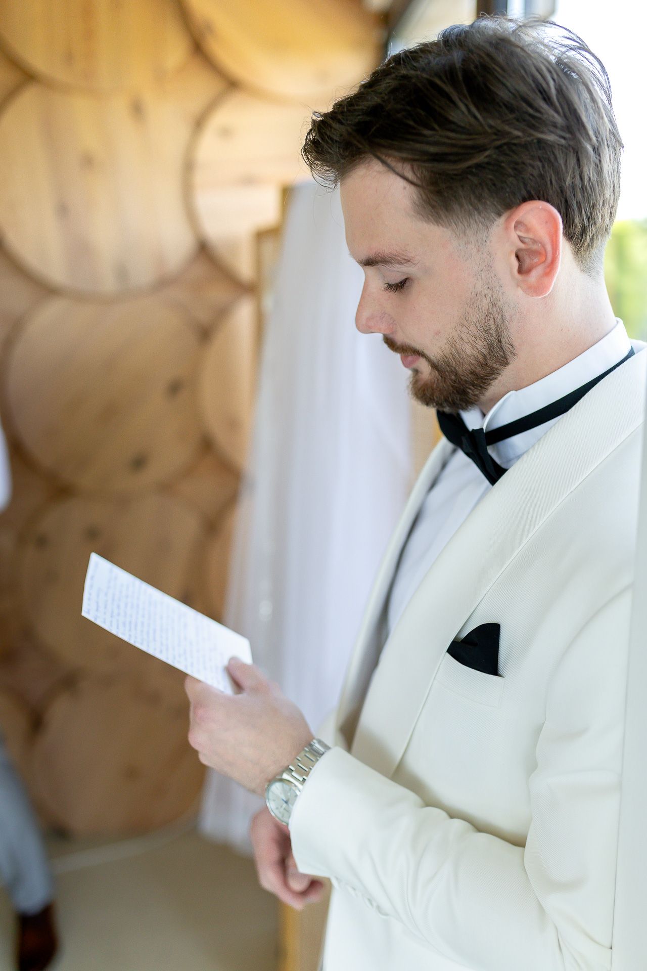A person in a white tuxedo reads from a paper in front of a textured wooden wall.