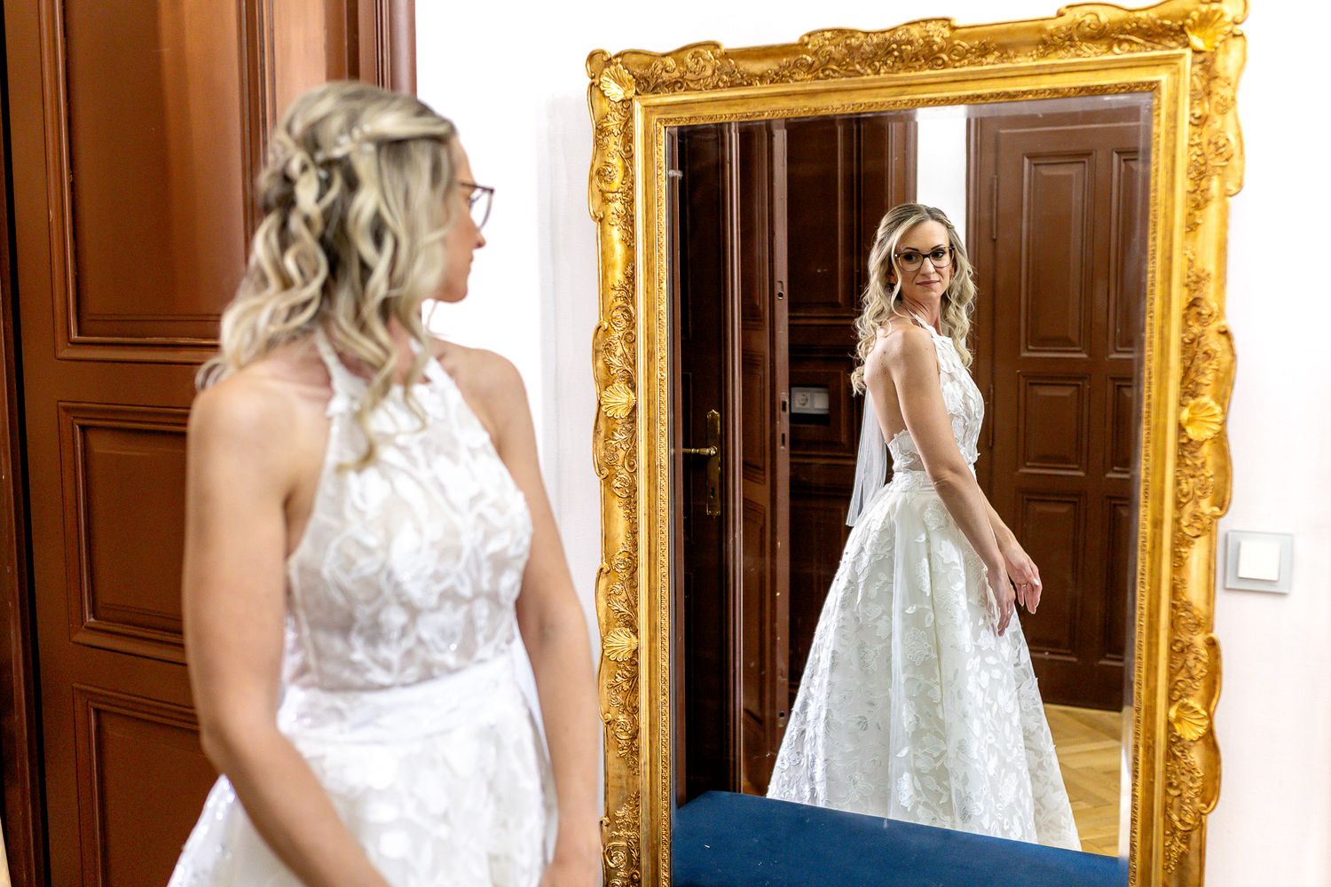 A bride in a white lace wedding dress stands in front of an ornate gold mirror in a bridal suite.