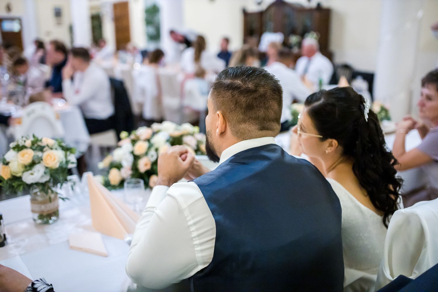 Elegant wedding reception table settings with white floral centerpieces and glowing candles in a bright venue.