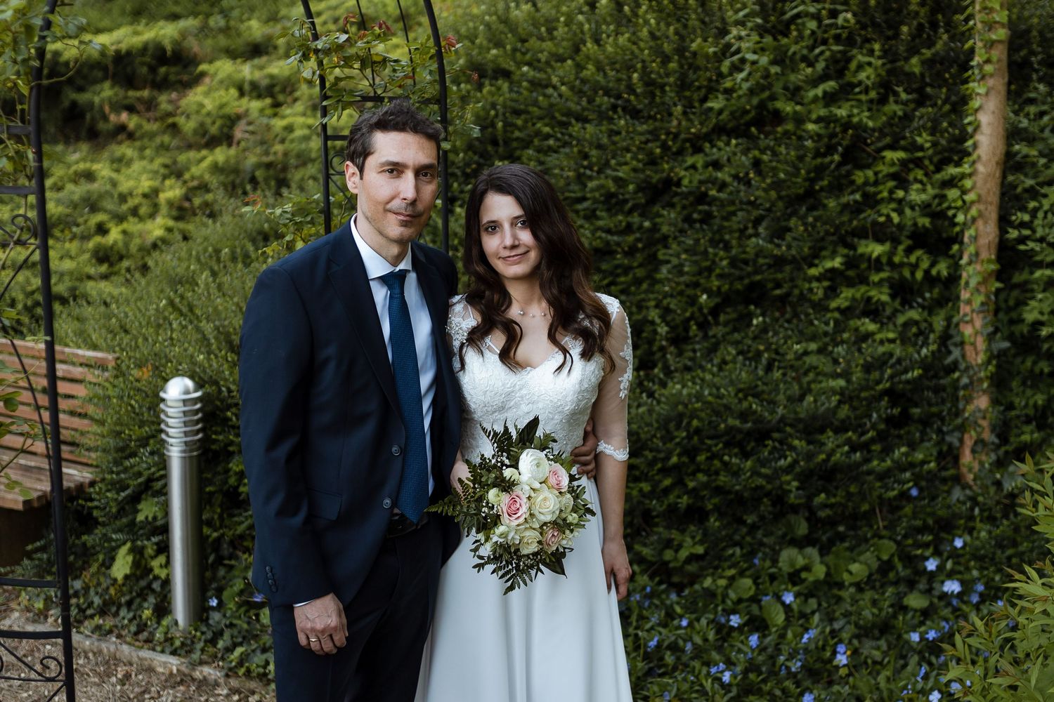 A couple stands together outdoors on their wedding day with greenery in the background and the bride holding a bouquet.