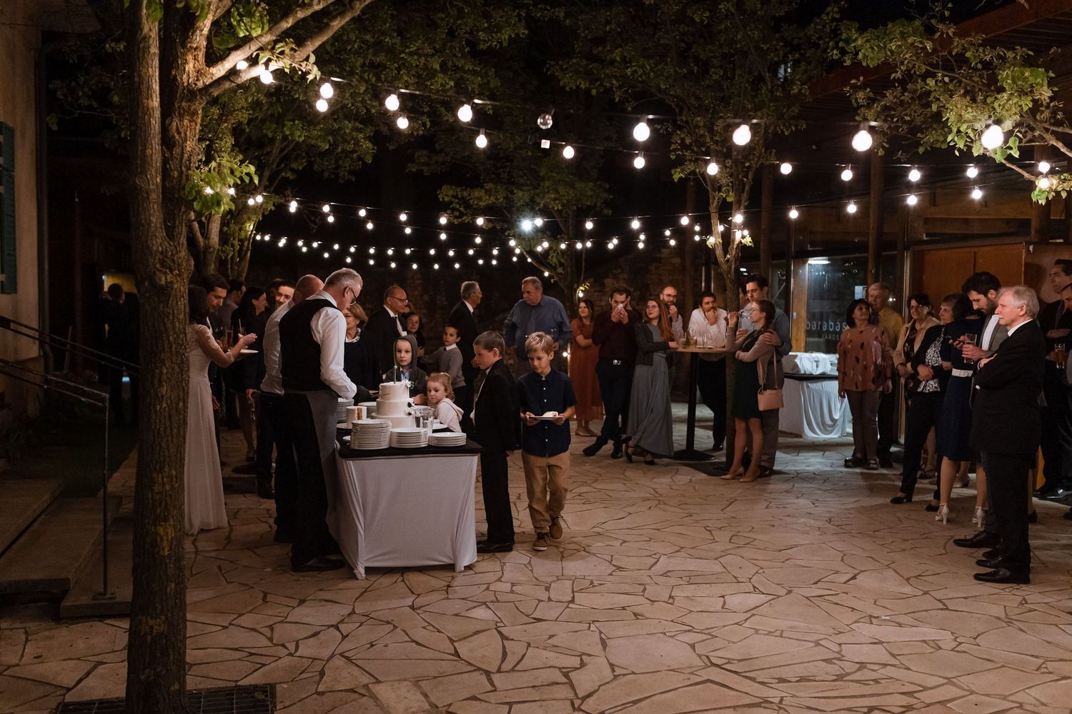 String lights illuminate an outdoor evening wedding reception with guests mingling under trees at dusk.