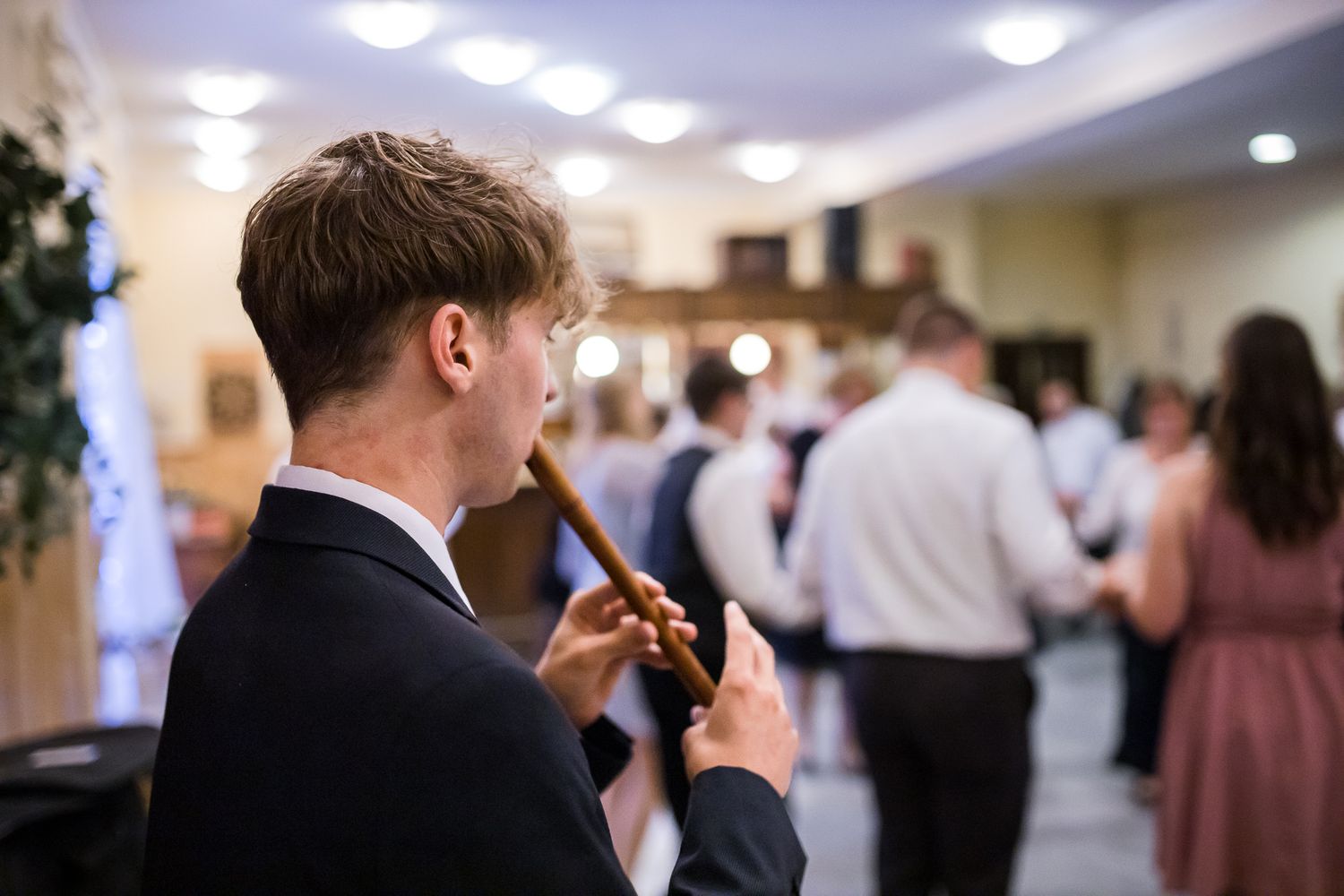 A person in a dark suit holds a phone at an indoor social gathering with other guests mingling in the background.